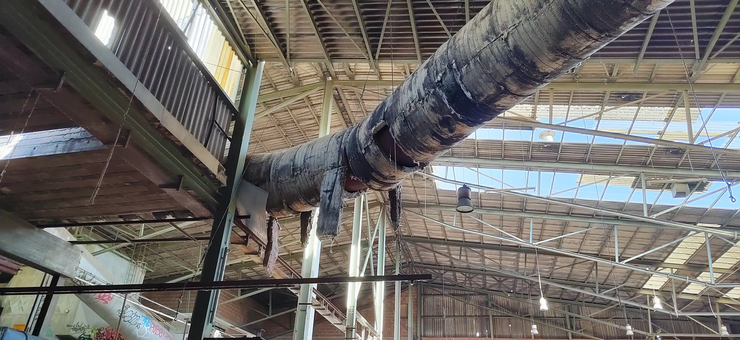 Brandschaden an einer großen Deckenleitung in einer Industriehalle, mit schwarzem Ruß und abgebrannten Stellen