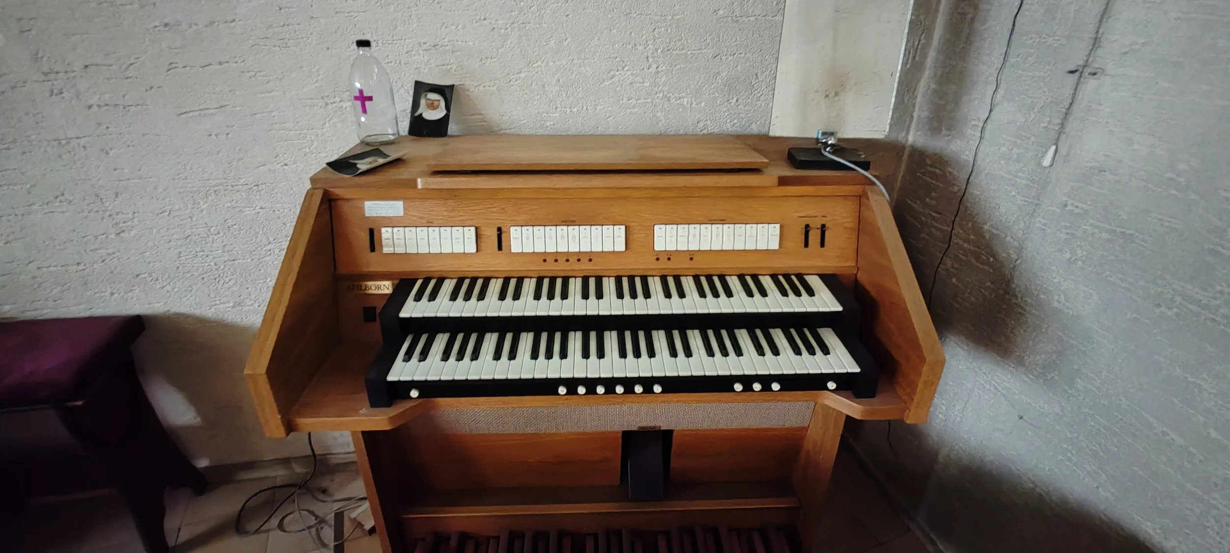 Ein Holz-Orgelpult mit mehreren Tasten, Reglern und Knöpfen. Auf dem oberen Teil des Instruments liegen einige Gegenstände, darunter eine Wasserflasche mit einem pinken Kreuz, ein kleines Foto und einige Papiere. Es ist an die Wand angeschlossen.