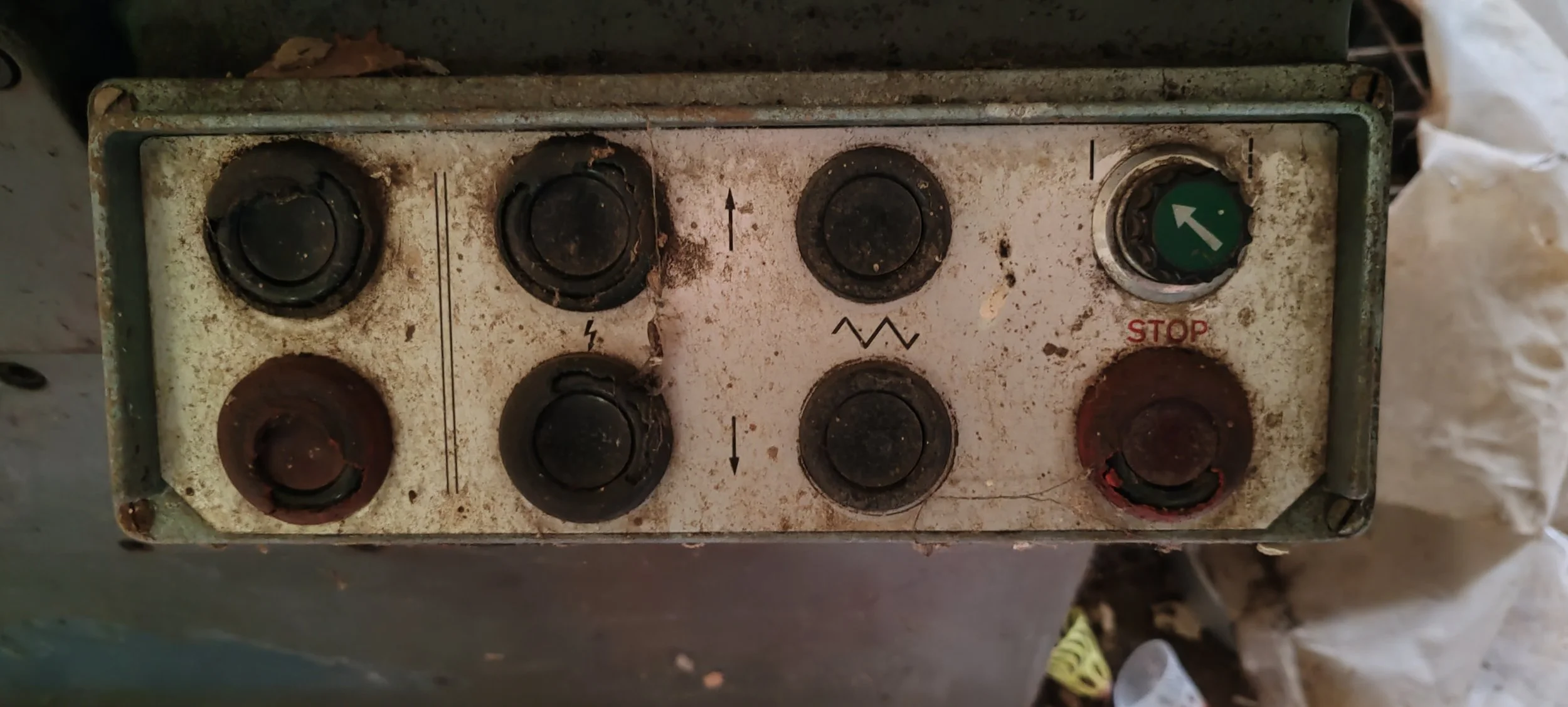 Alter, verrosteter Steuerkasten mit sechs Knöpfen, einigen Markierungen, und einem roten Stop-Button, alles verschmutzt und abgenutzt.