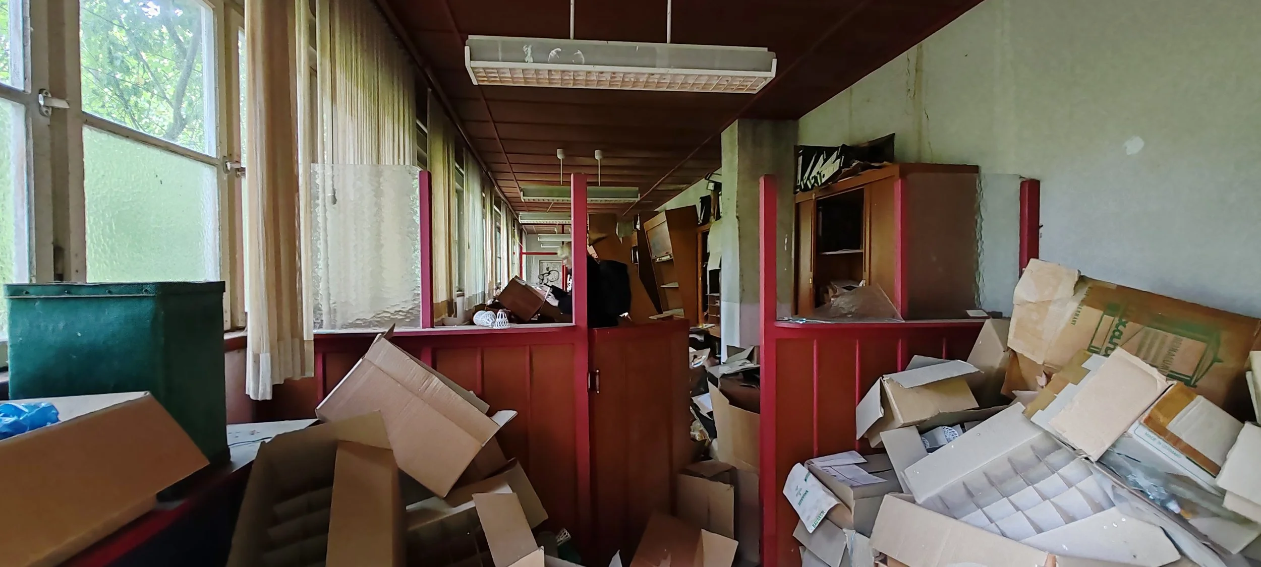 Verlassenes und chaotisches Zimmer mit durchwühlten Schreibtischen, Cartons und verstreutem Papier. Fenster an der linken Seite lässt Tageslicht herein, einige Möbel sind beschädigt oder umgestürzt.