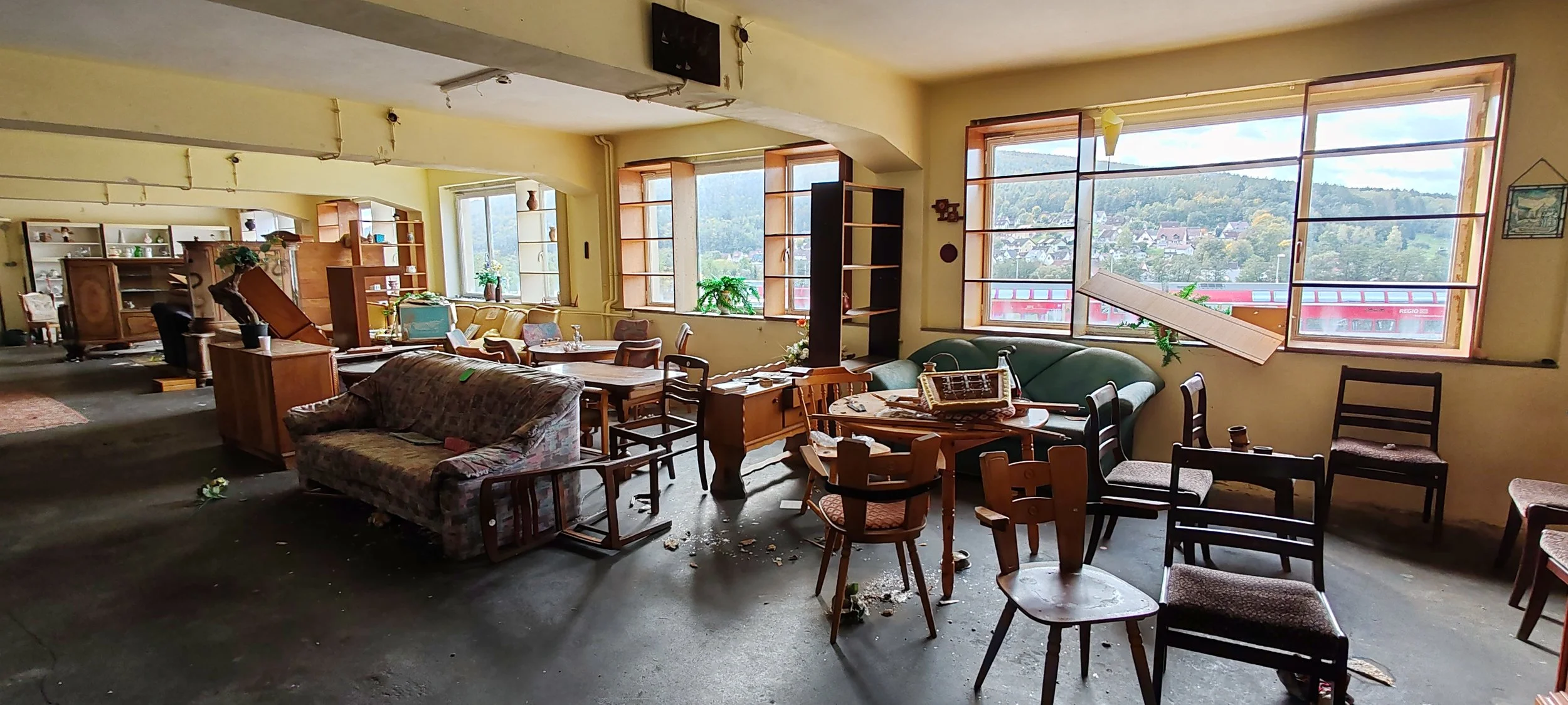 Verlassene und chaotische Wohnzimmer mit umgestürzten Möbeln, verstreuten Gegenständen und offenstehenden Fenstern, die auf eine Einbruch- oder Katastrophenszene hinweisen.