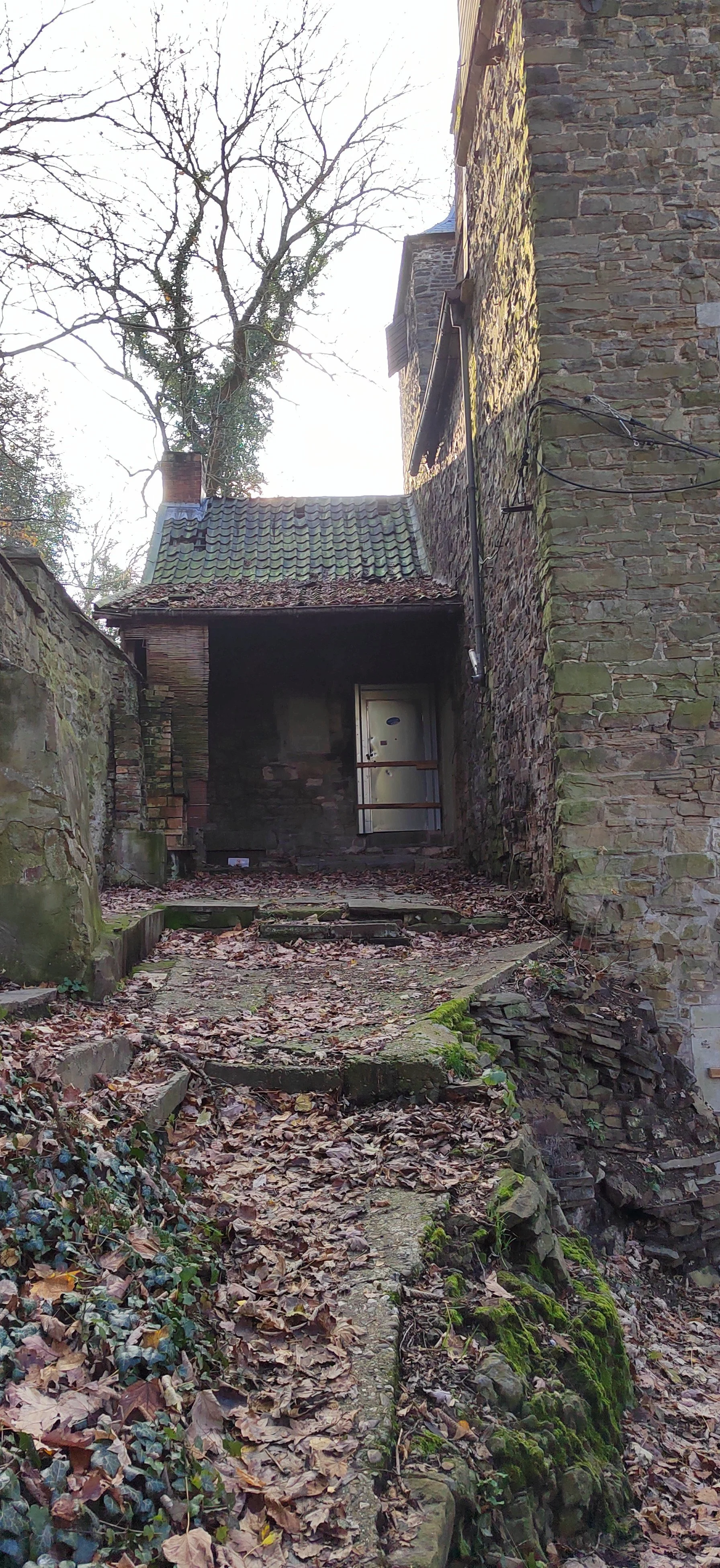 Ein altes, steinernes Gebäude mit einer steilen Dachterrasse, umgeben von Laubbäumen und einem mit Laub bedeckten, unebenen Weg im Freien.
