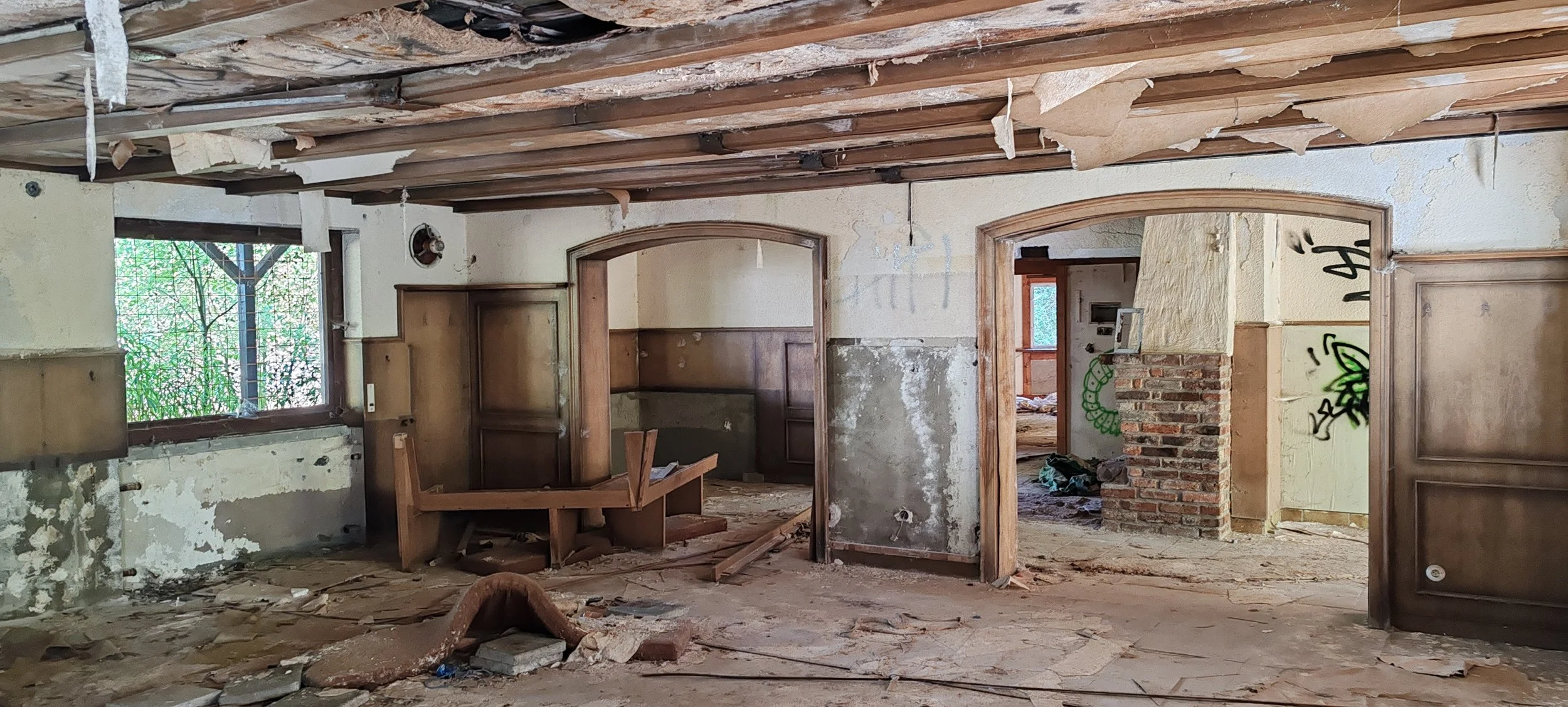 Innenraum eines zerstörten Hauses mit beschädigter Decke, zerbrochenen Wänden, einem offenen Kamin und bemalter Wand. Das Haus ist verlassen und offensichtlich renovierungsbedürftig.