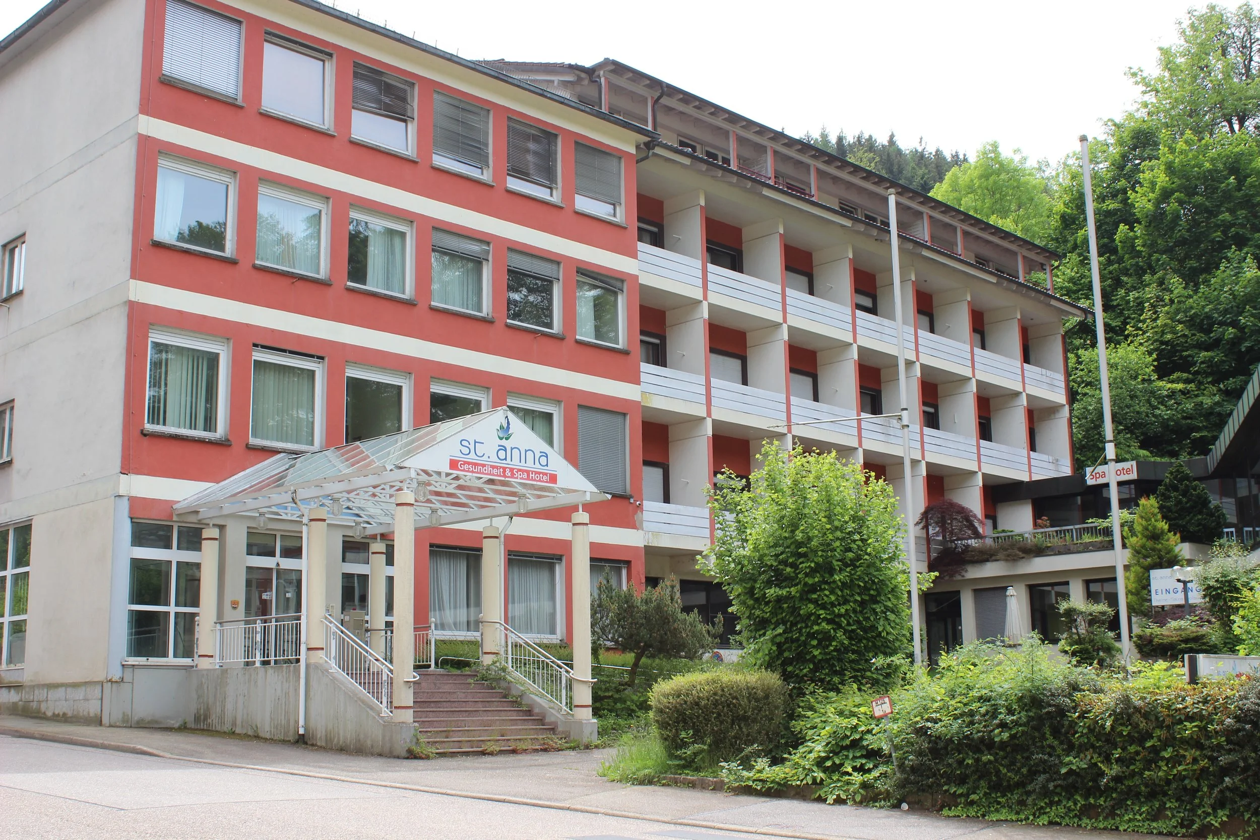 Mehrstöckiges Hotelgebäude mit roten und weißen Fassaden, Eingangsbereich mit Treppe und Beschilderung »st. anna« und »Gesundheit & Spa Hotel«, umgeben von Bäumen und Garten. Hinter dem Gebäude sind grüne Hügel.