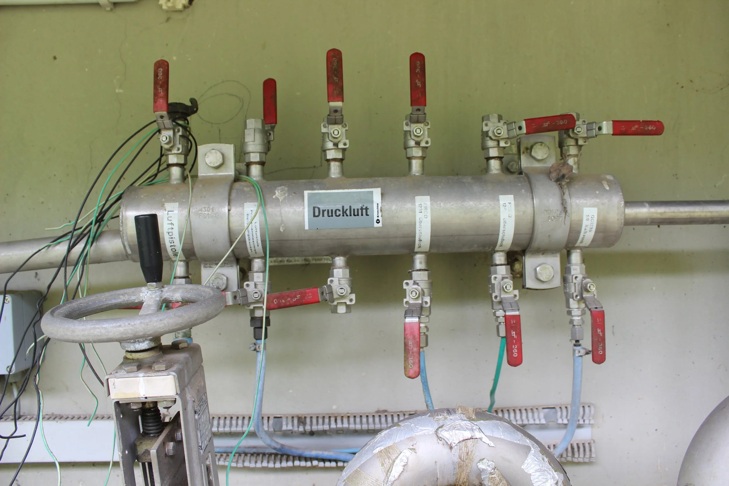 Druckluftsystem mit mehreren roten Absperrventilen, verbunden mit Rohren, an einer grauen Wand montiert.