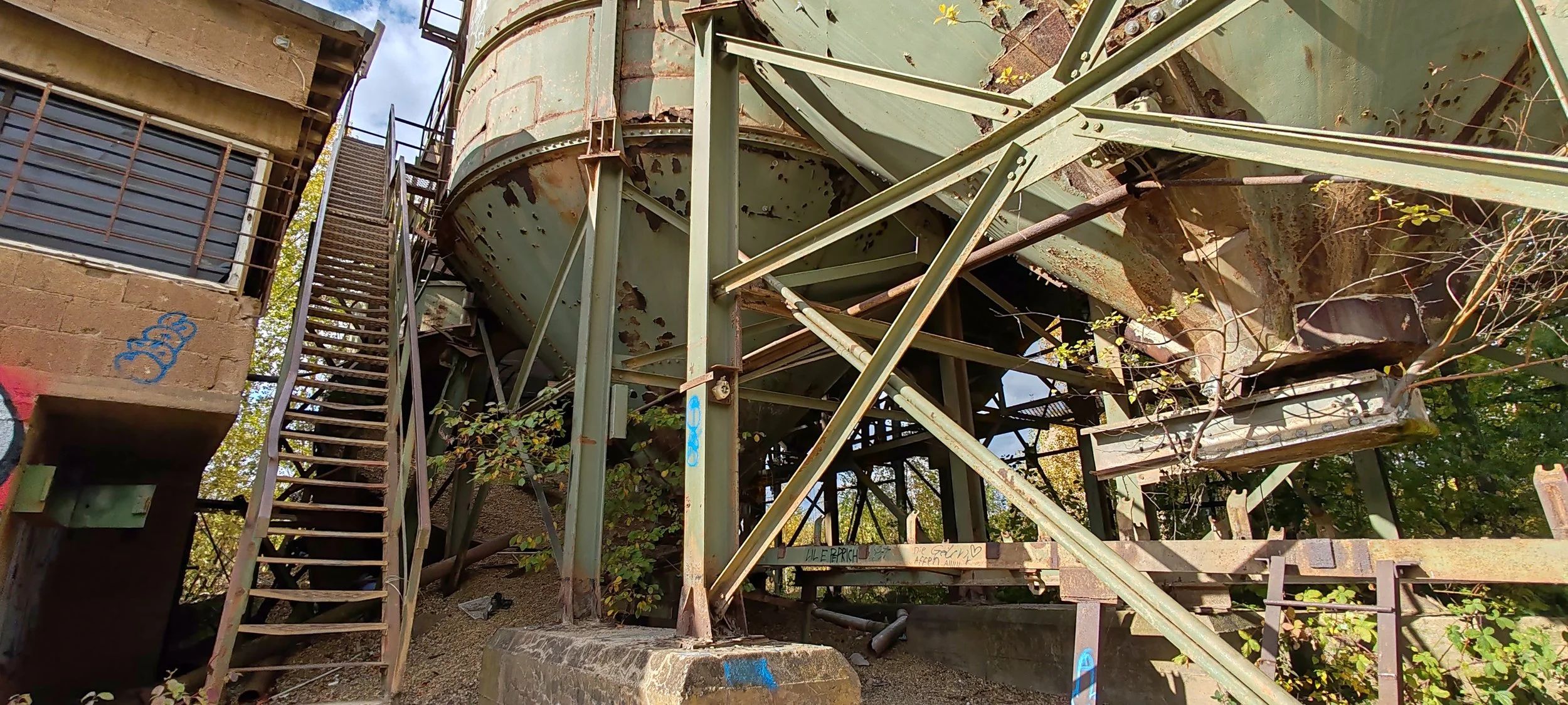 Ein verrosteter, grüner Wasserbehälter auf einem alten Industriegelände, umgeben von rostigen Metallstrukturen, Treppen und Graffiti an einer Betonmauer.