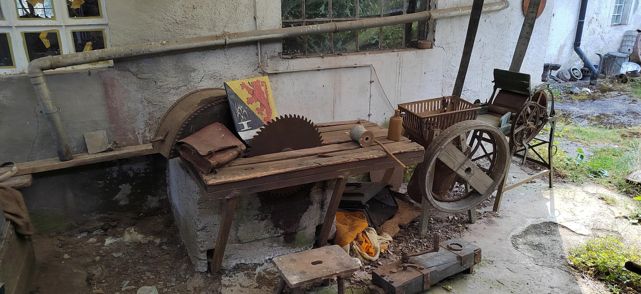 Alte Werkzeuge und Maschinen im Außenbereich, inklusive Säge, Kisten, Pumpe und anderen Holzteilen.