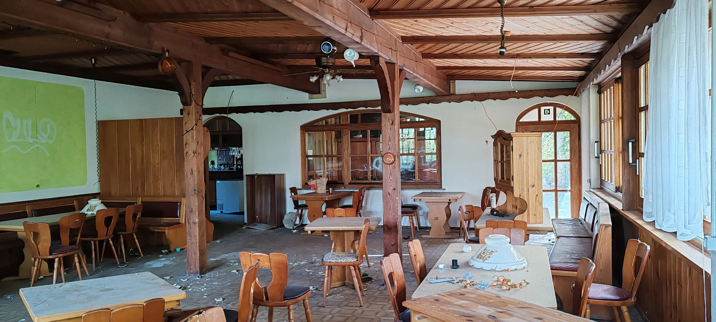 Innenansicht eines verfallenen Restaurants oder Cafés mit Holzdekoration, mehrere Tische und Stühle, einige Gegenstände auf den Tischen, zerbrochene Kaffeetasse und überall verstreute Trümmer.