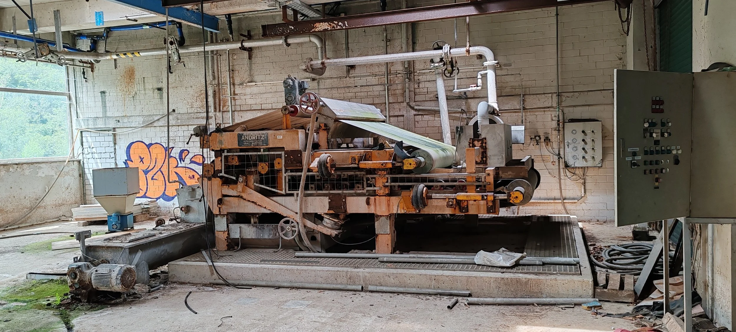Verrostete industrielle Maschine in einer verfallenen Fabrikhalle mit Graffiti an der Wand im Hintergrund.