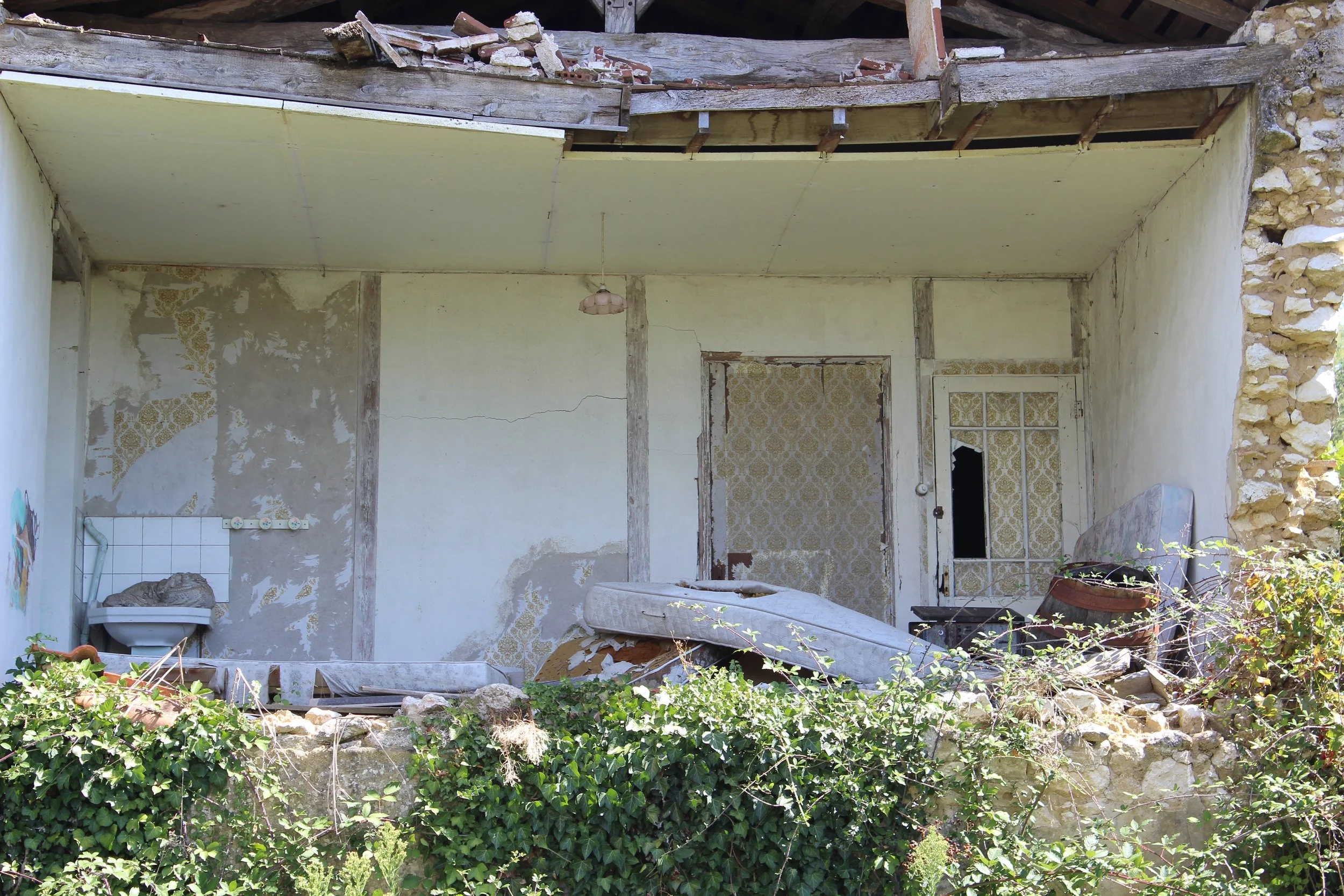 Verlassene und zerfallene Hausfassade mit beschädigten Wänden, zerbrochenen Fenstern und Möbeln im Inneren, überwuchert von Pflanzen.