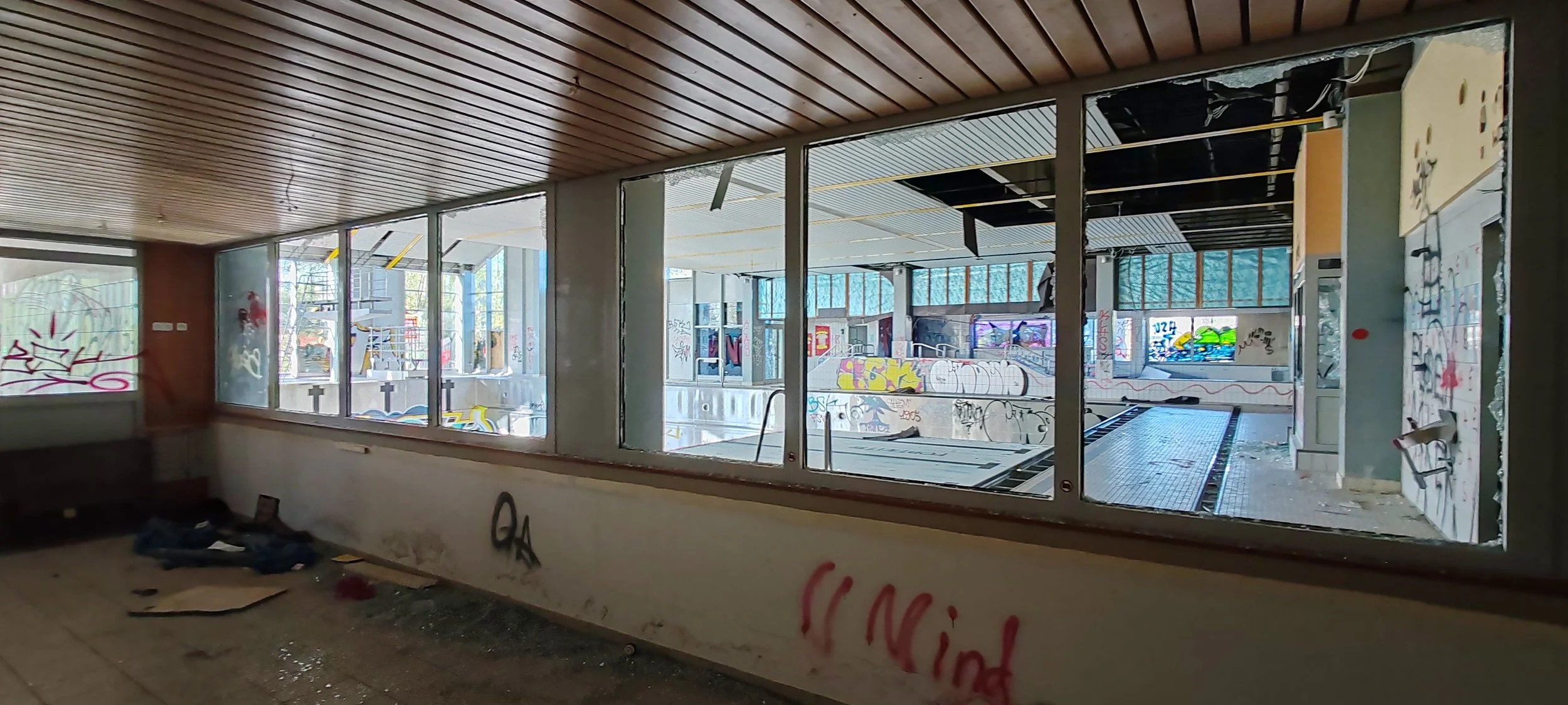 Verlassener, vandalismusbeschädigter Swimmingpool in einem Gebäude mit Graffiti an den Wänden, kaputtem Glas und verfallenem Zustand.