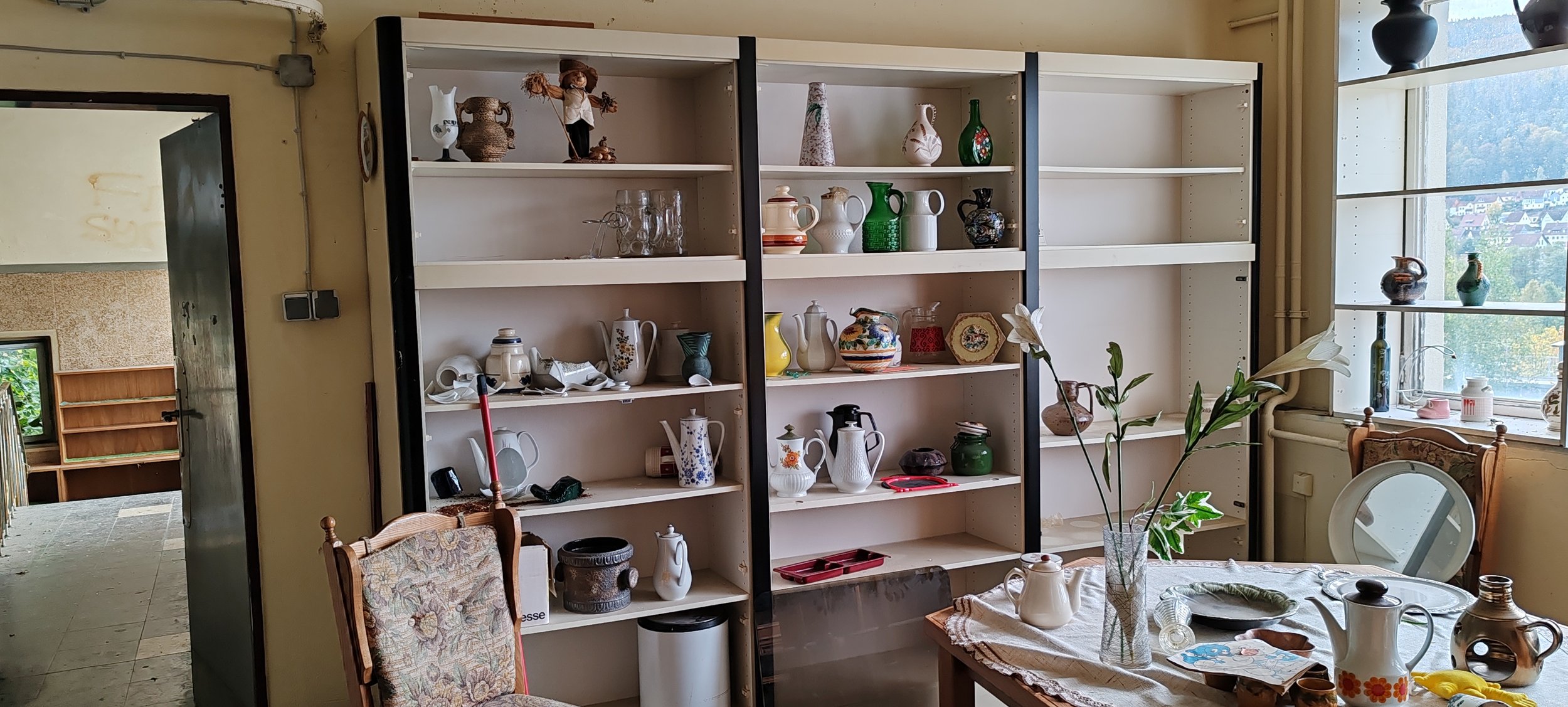 Bunte Keramikteller und Vasen auf leeren weißen Regalen in einem hellen Raum neben einem Fenster, das einen Blick auf die Stadt bietet.