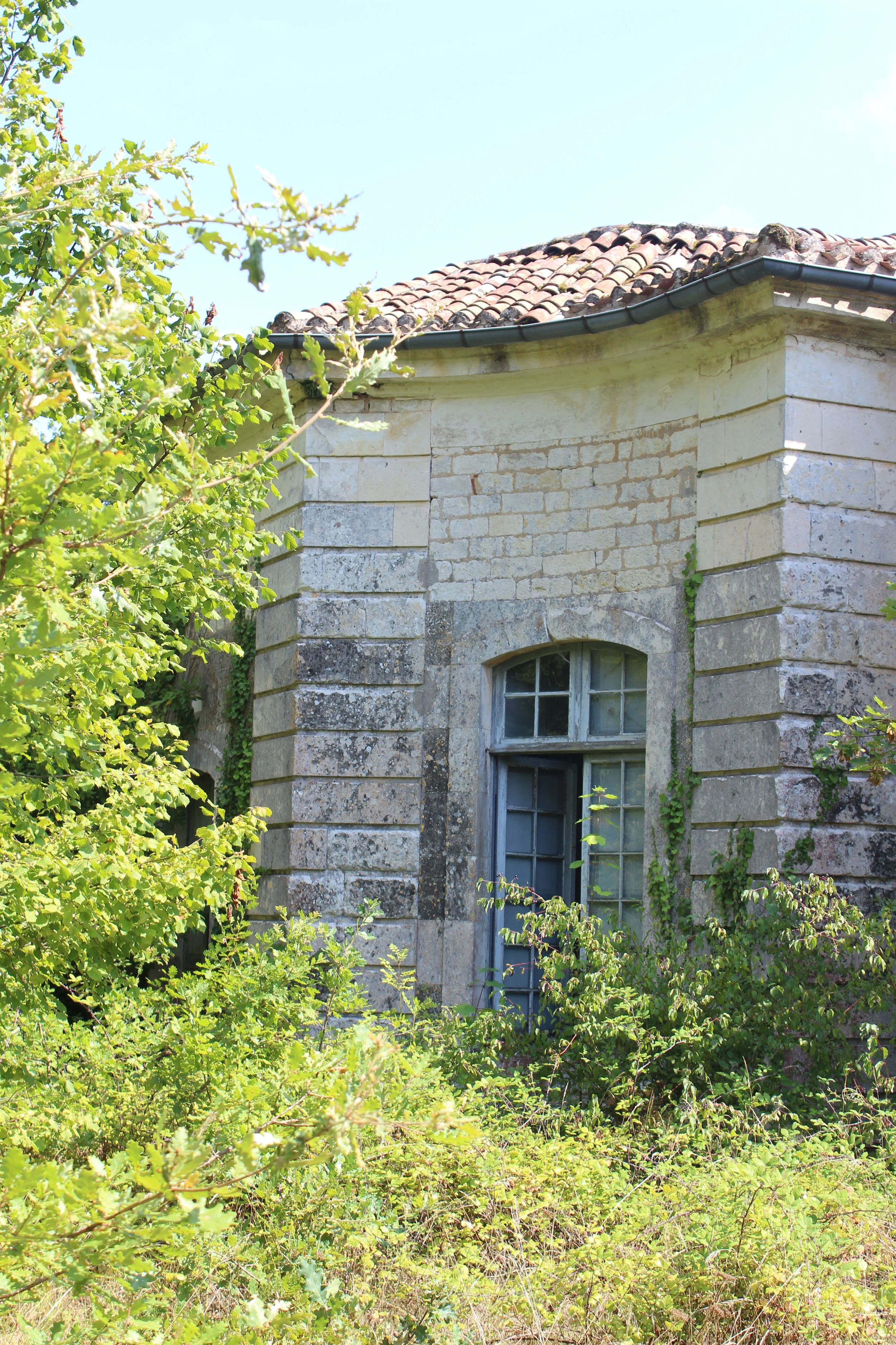 Ein altes Steinhaus mit einer runden Bauweise, einer kleinen, vergitterten Fenster und einem Ziegeldach, umgeben von grünen Pflanzen und Bäumen.