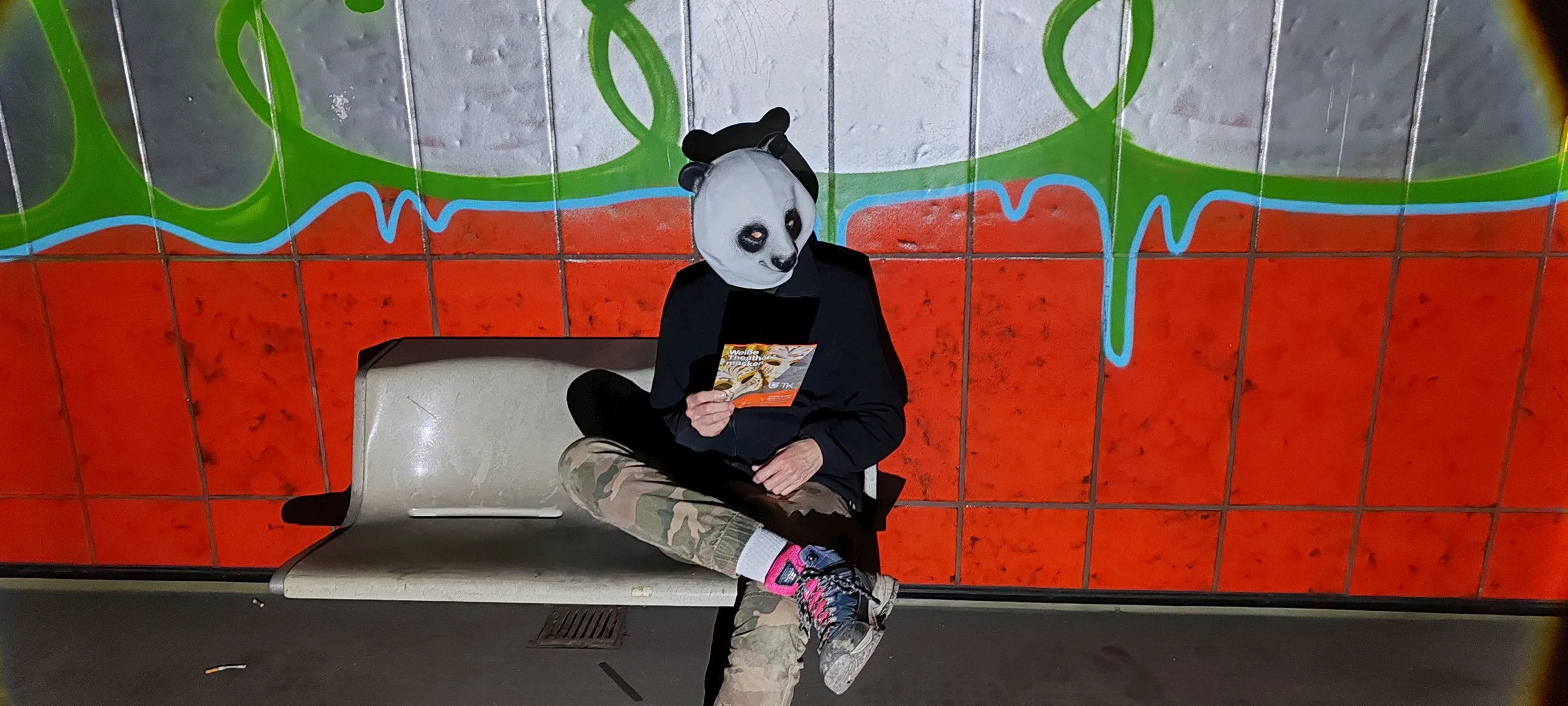 Person mit Panda-Maske sitzt auf einer Bank, liest eine Broschüre, trägt Camouflage-Hose, pinke Socken und Sportschuhe, im Hintergrund eine bunte Wand.