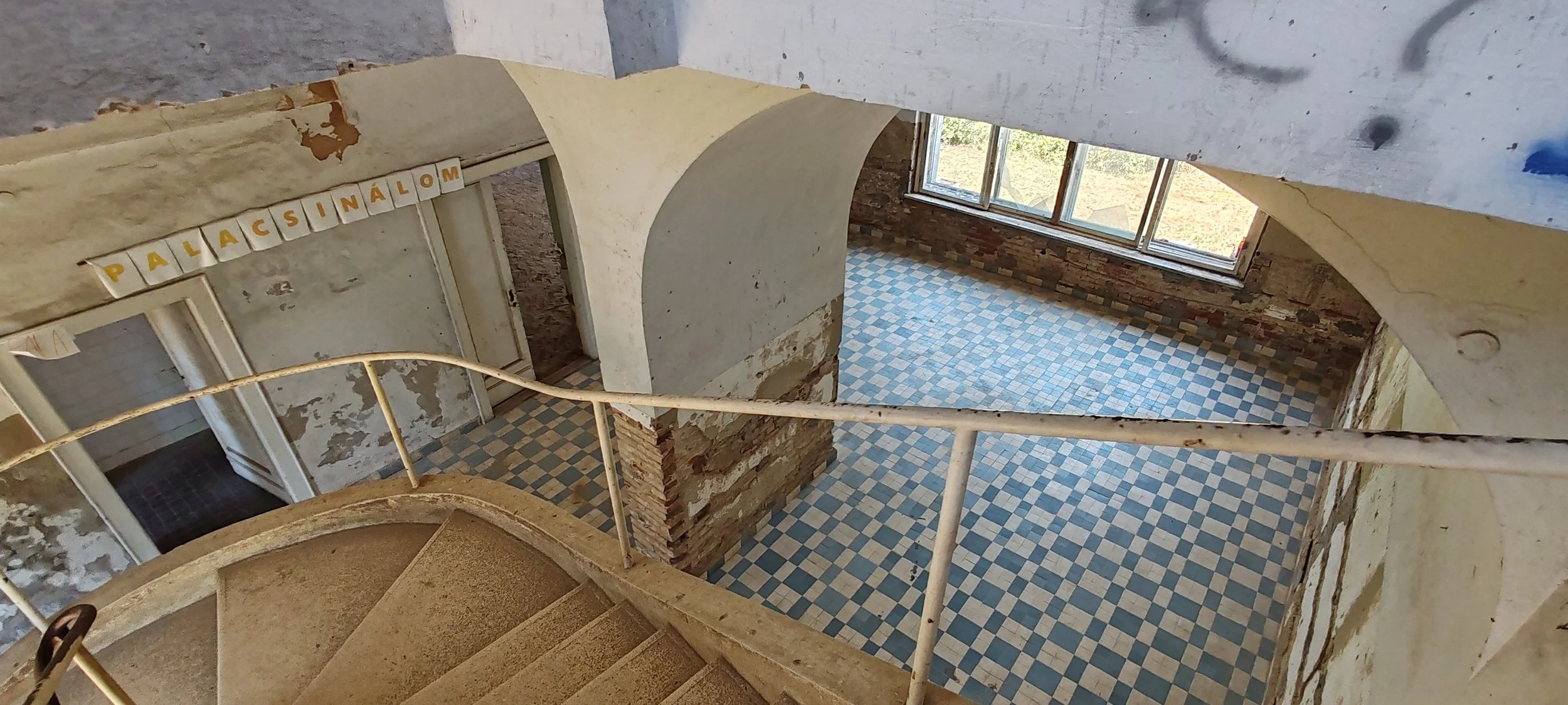 Innenansicht eines alten Gebäudes mit welliger Treppe, blau-weißen Fliesen, großen Fenstern und abblätternder Wandfarbe. Es sind Buchstaben an der Wand zu sehen, die das Wort 'PALÄCSINÄLÖM' bilden.