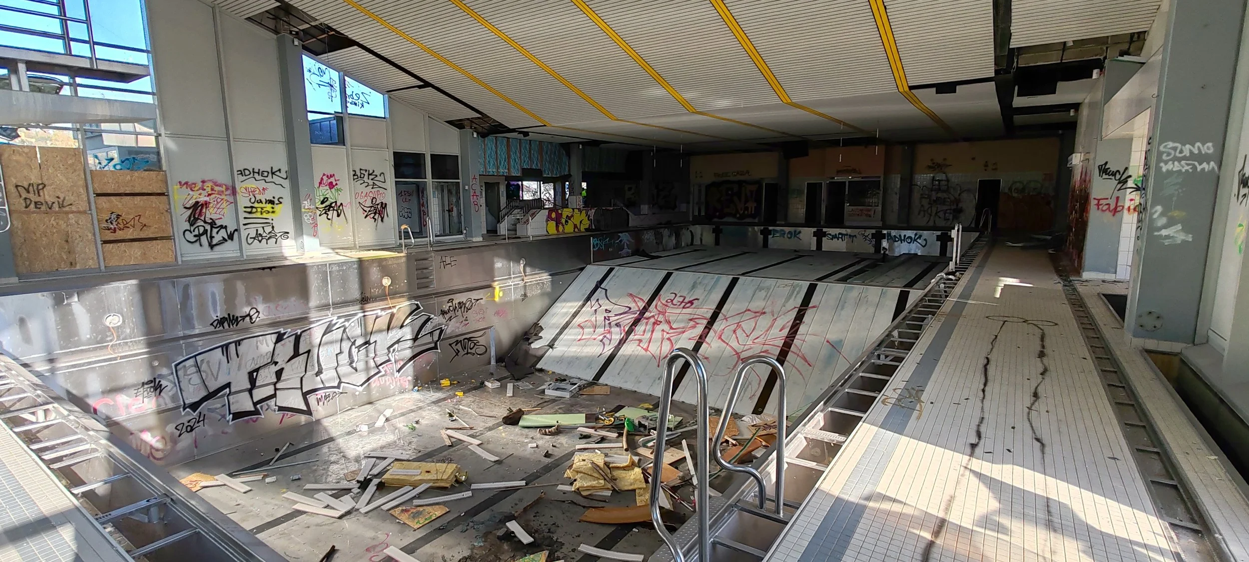 Verlassenes, verwahrlostes Schwimmbad mit Graffiti, beschädigten Fliesen und Müll im Wasserbecken.