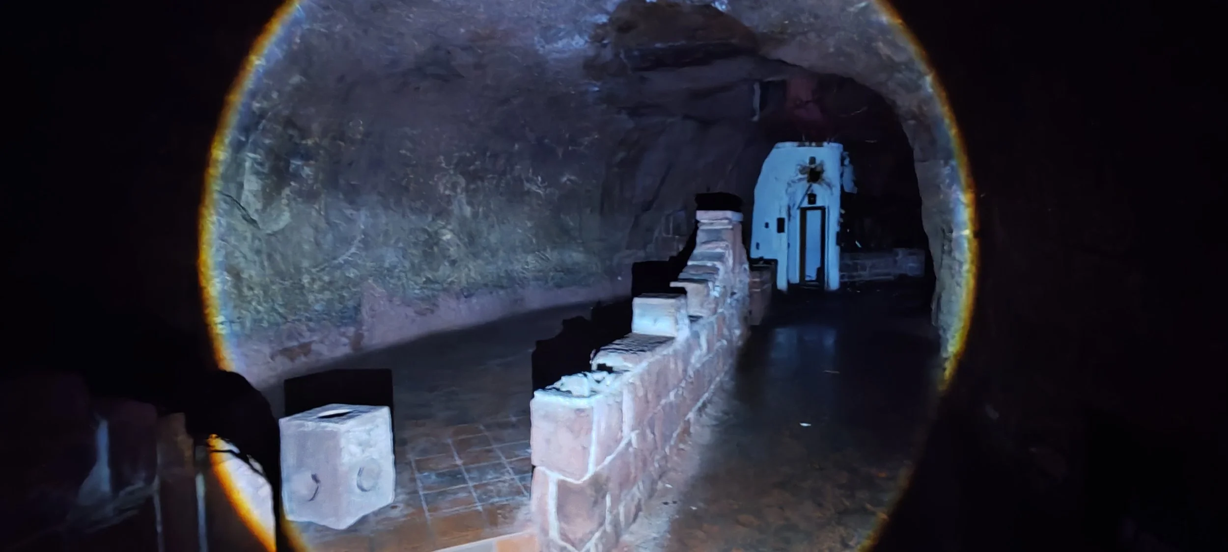 Blick durch ein Teleskop in eine Höhle mit Steinbänken und alten Geräten, dunkle Atmosphäre, steinige Wänden, dunkler Boden.