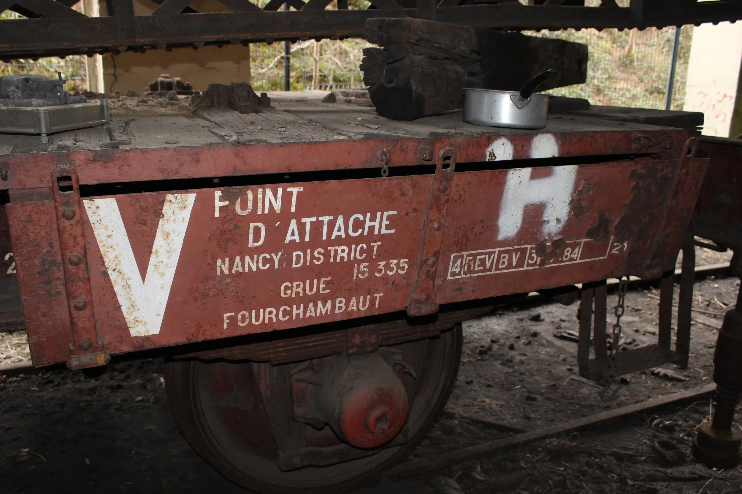 Alter roter Eisenbahnwagen mit weißen Aufschriften, der auf Schienen steht, mit einem Kochtopf auf der Plattform. Im Hintergrund ist eine Holzwand und Natur zu sehen.