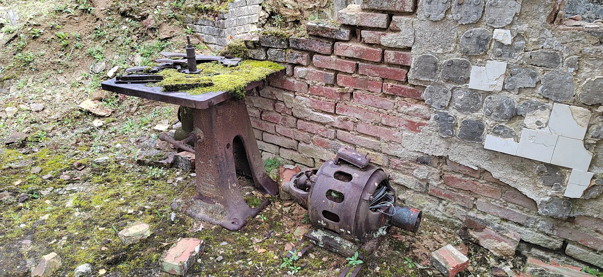 Ein alter, verrosteter Schneidbrenner auf einer Steinbasis vor einer Mauer mit kaputten Fliesen und Mauerwerk, mit Moos und Pflanzen auf dem Boden.