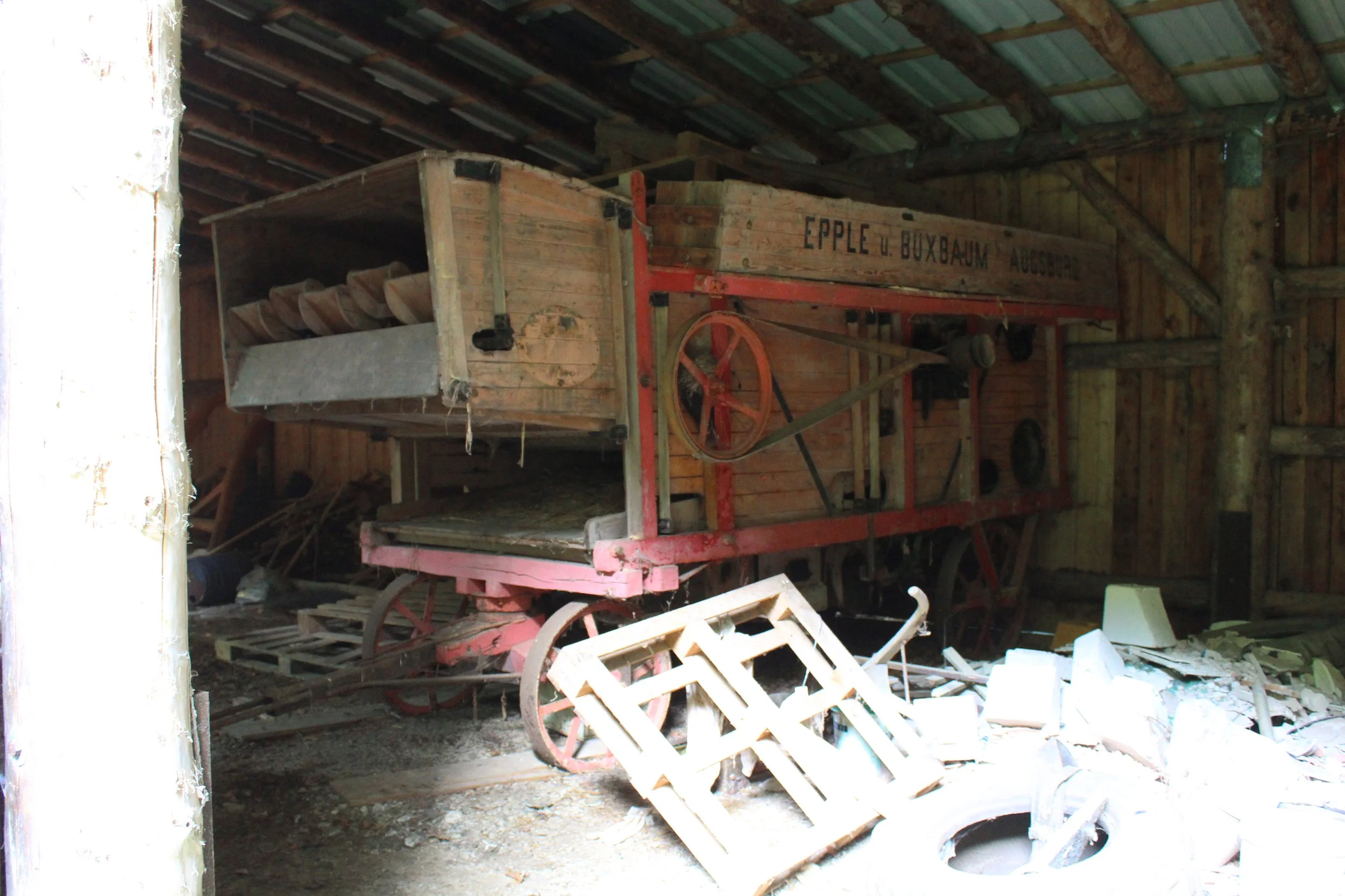 Ein alter, verstaubter nostalgischer Holz-Landwirtschaftswagen mit roten Metallteilen, im Inneren eines verlassenen Scheunenraums, umgeben von Baumaterialien und Schutt.
