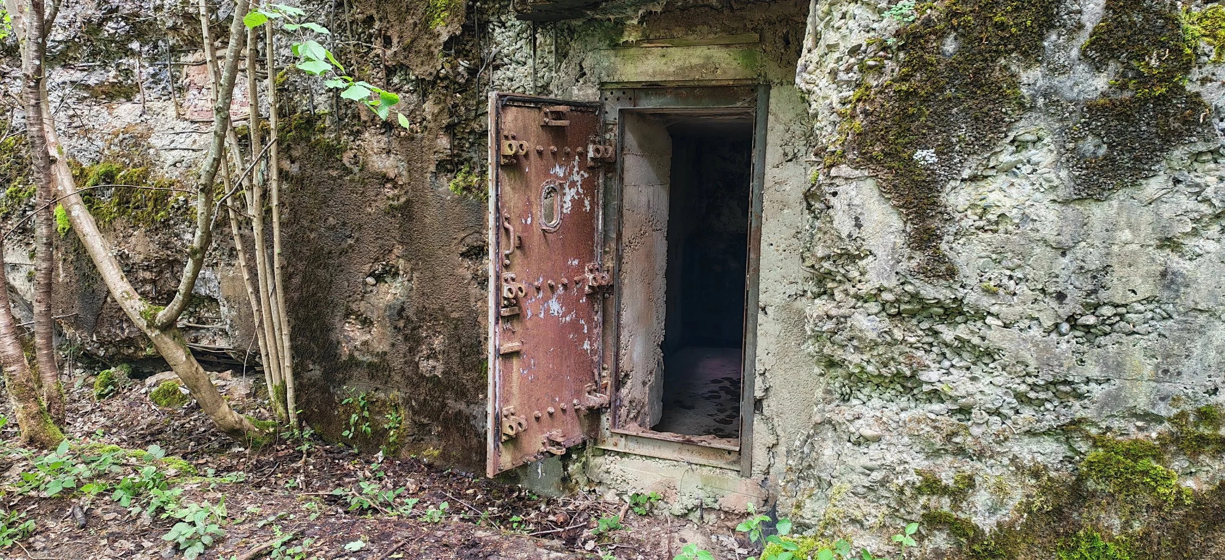 Ein alter, rostiger Bunker mit offenem Eingang, umgeben von natürlicher Vegetation und Steinen.