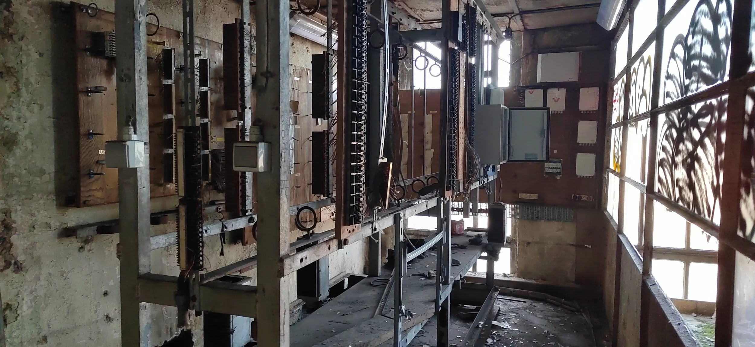 Veraltete, nicht funktionierende komplexe elektronische Schaltanlage mit Kabeln, Steuereinheiten und Metallrahmen in einem alten, verfallenen Gebäude mit großen Fenstern, durch die Licht fällt.