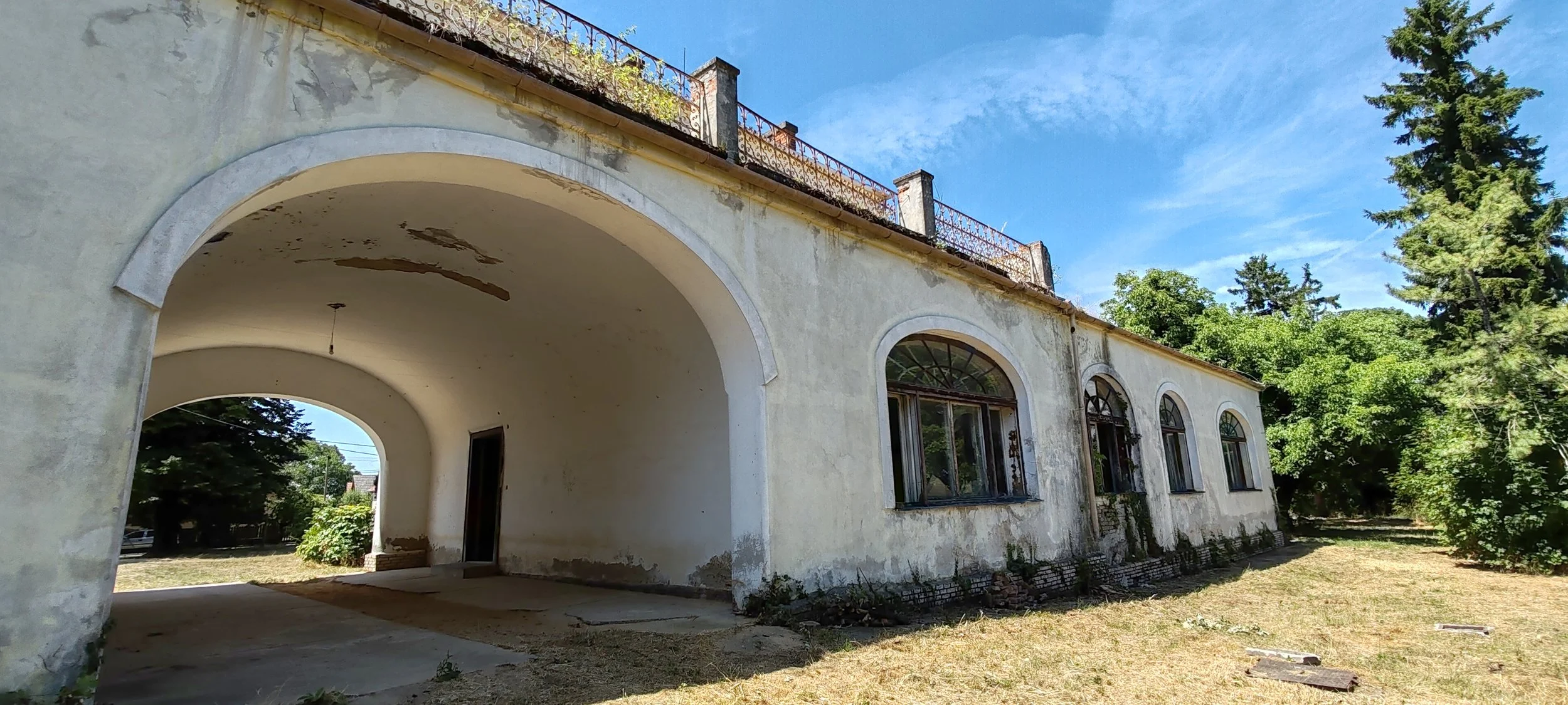 Verfallenes, altes Gebäude mit großen, arched Fenstern und einem offenen Durchgang im Vordergrund, umgeben von Bäumen, bei sonnigem Himmel.