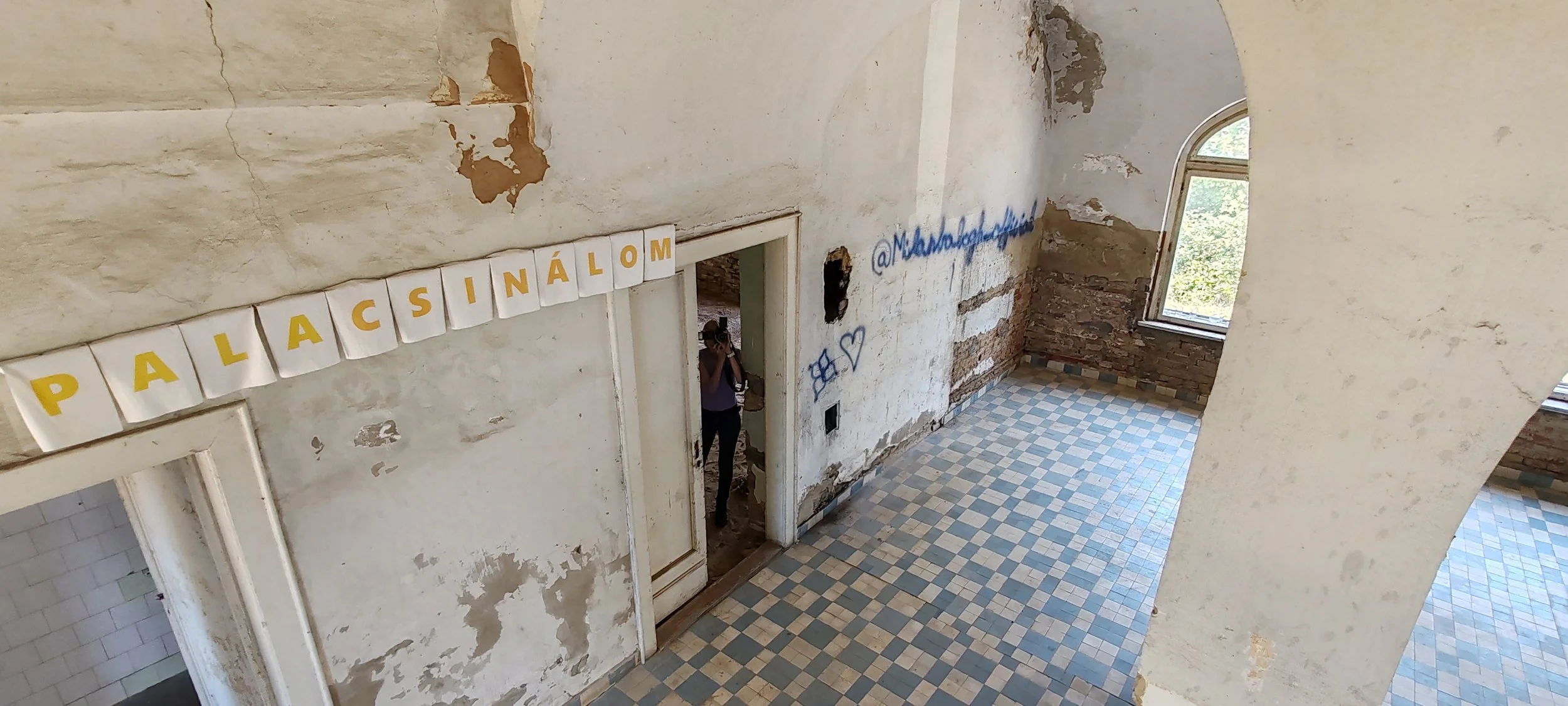 Innenansicht eines alten, verfallenen Hauses mit verputzten Wänden, einem großen Fenster, und einem Fliesenboden in Blau- und Cremetönen. An der Wand hängt eine Banner mit weißen Karten, auf denen in gelbfarbenen Buchstaben 'PALACINÁLOM' geschrieben 