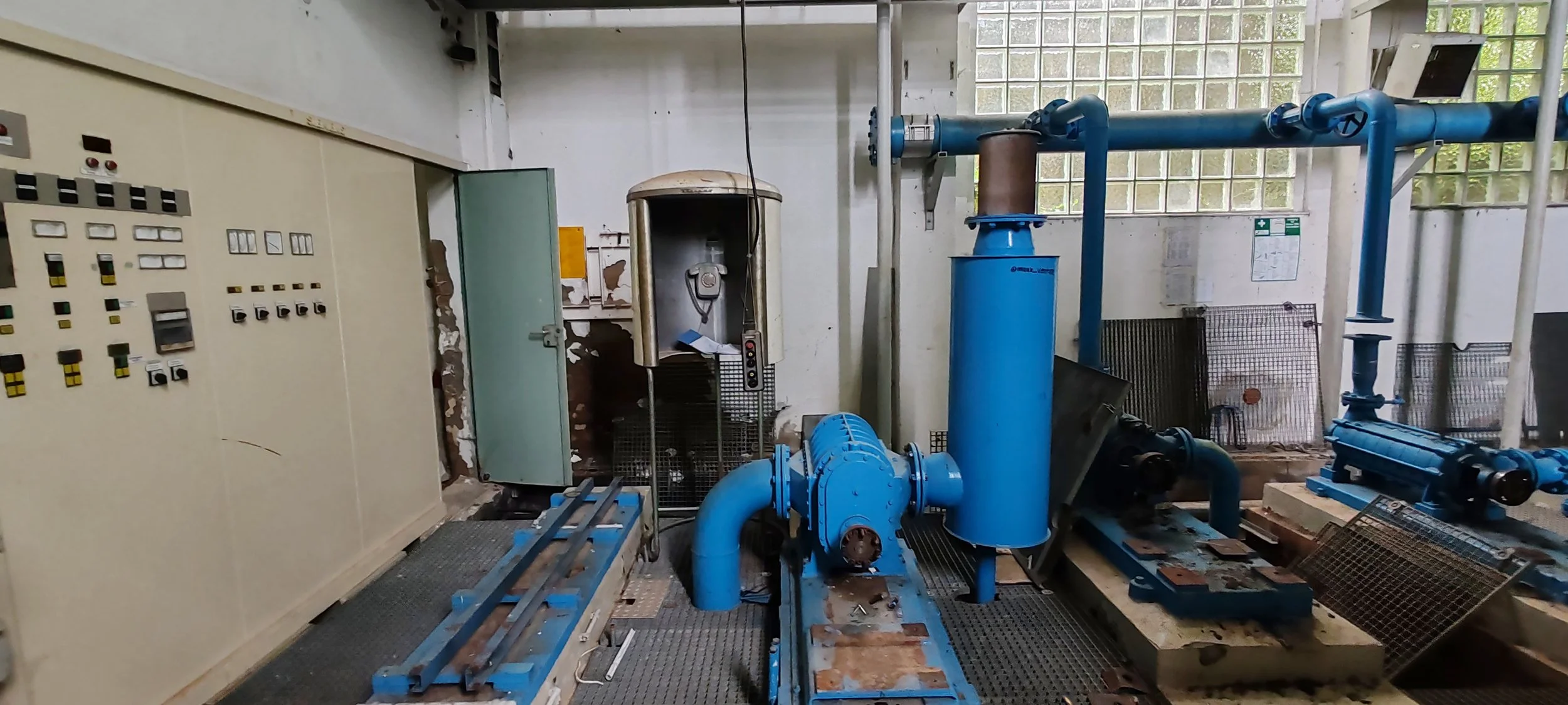Industrielle Anlage mit blauen Rohren, Pumpen, Steuerungskästchen und einer alten Telefonzelle in einem Fabrikraum.