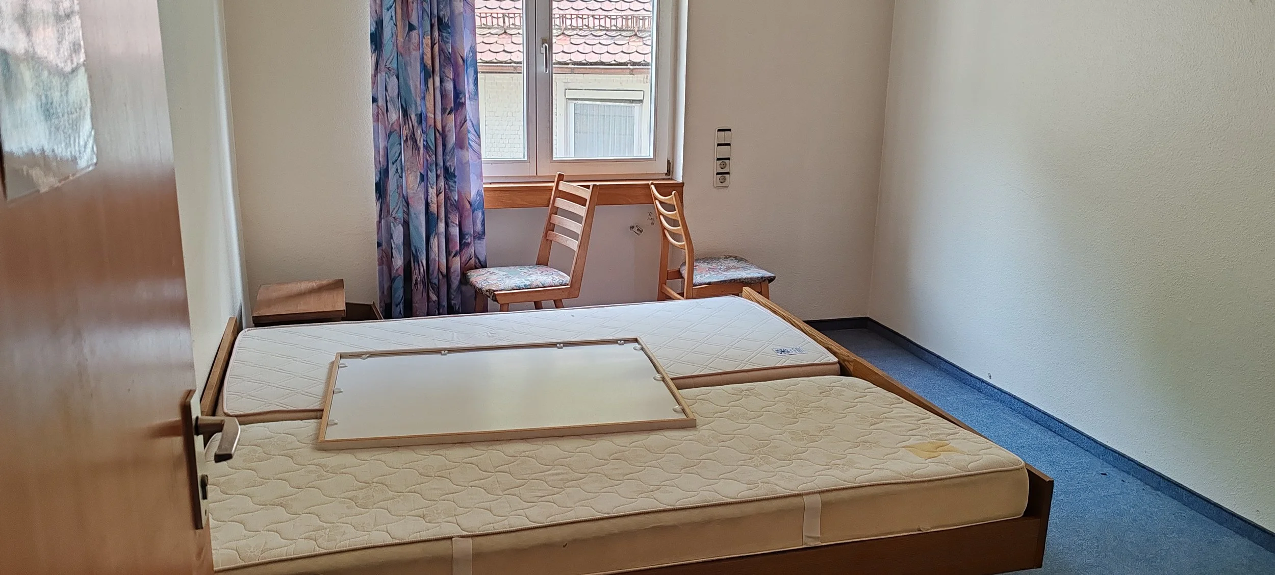 Ein leeres Schlafzimmer mit einem Doppelbett, zwei Holzstühlen vor einem Fenster mit bunten Vorhängen, und einem kleinen Nachttisch.