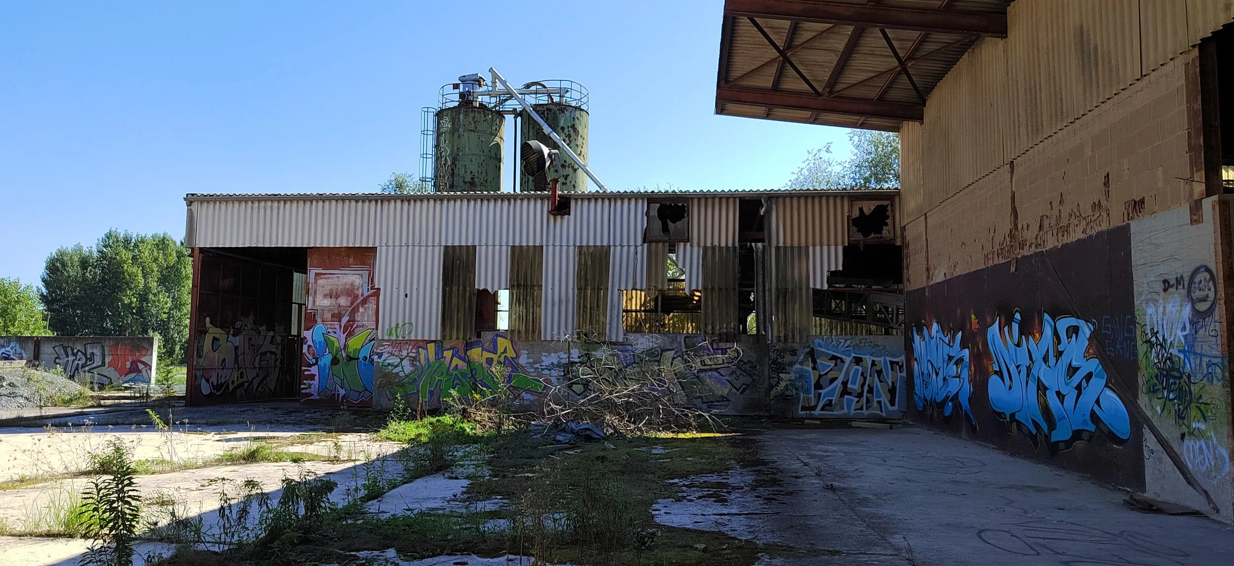 Verfallenes, graffiti-überwachtes Fabrikgebäude mit grünen Silos im Hintergrund.