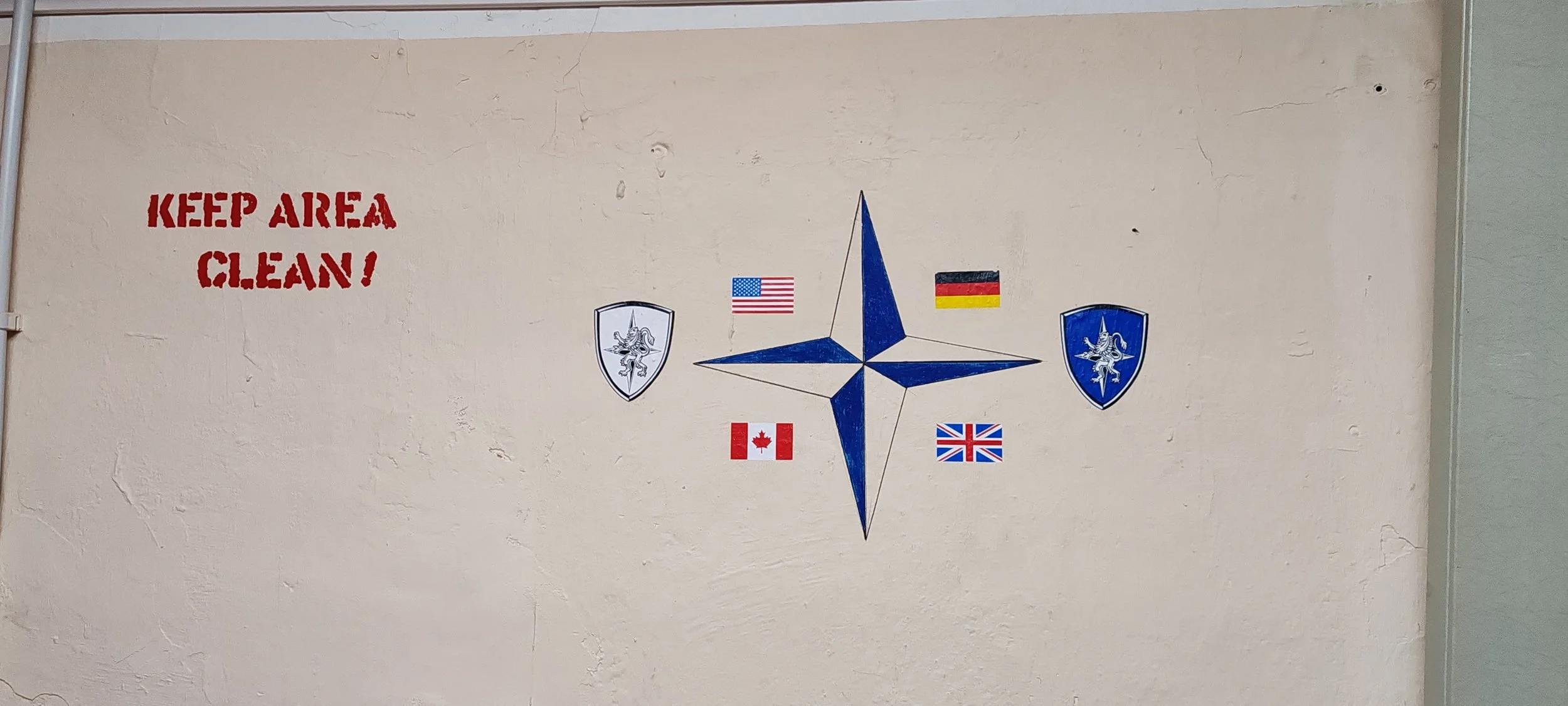 Wand mit der Aufschrift "Keep Area Clean!" in roter Schrift, ein blauer Kompass mit Flaggen der USA, Kanada, Großbritannien, Deutschland, Frankreich, und den Niederlanden, sowie zwei Wappen mit Einhornmotiven auf weißen und blauen Schildern.