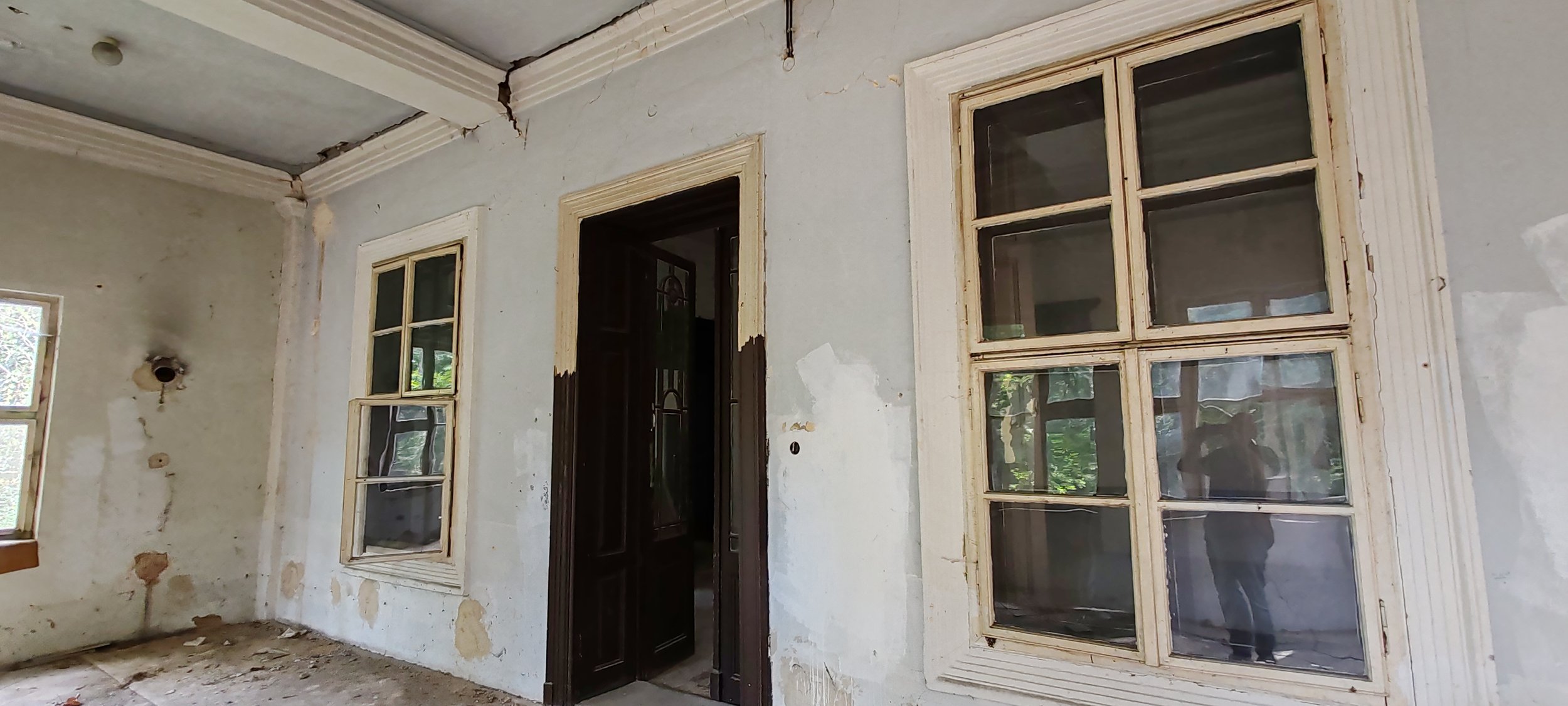 Ein altes, verfallenes Zimmer mit drei unvollständigen Fenstern und einem offenen Türrahmen vor einer beschädigten Wand. Die Wand und der Boden sind beschädigt und veraltet.