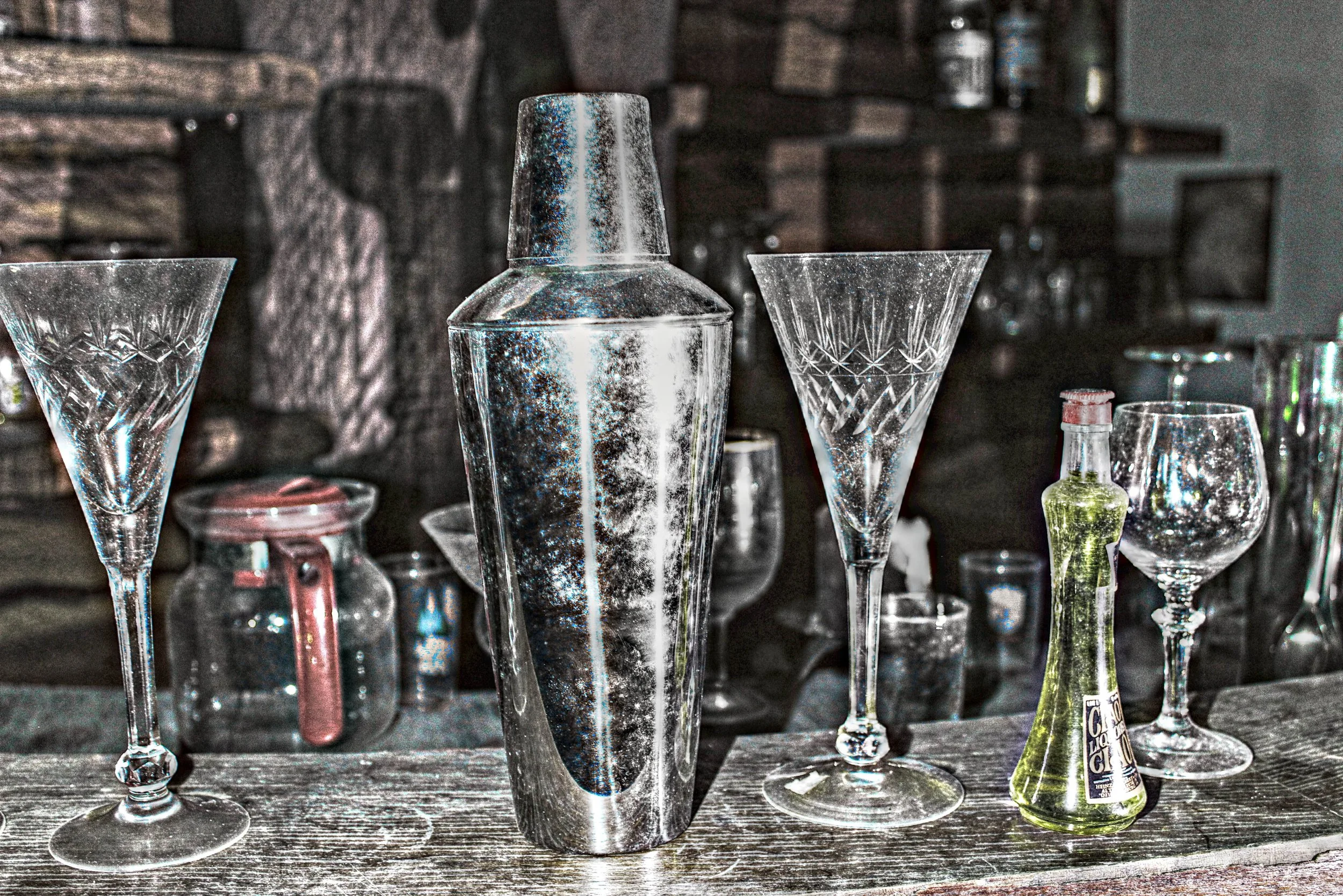 Verschiedene leere Bar- oder Cocktailgläser auf einem Tisch, darunter eine grüne Flasche.