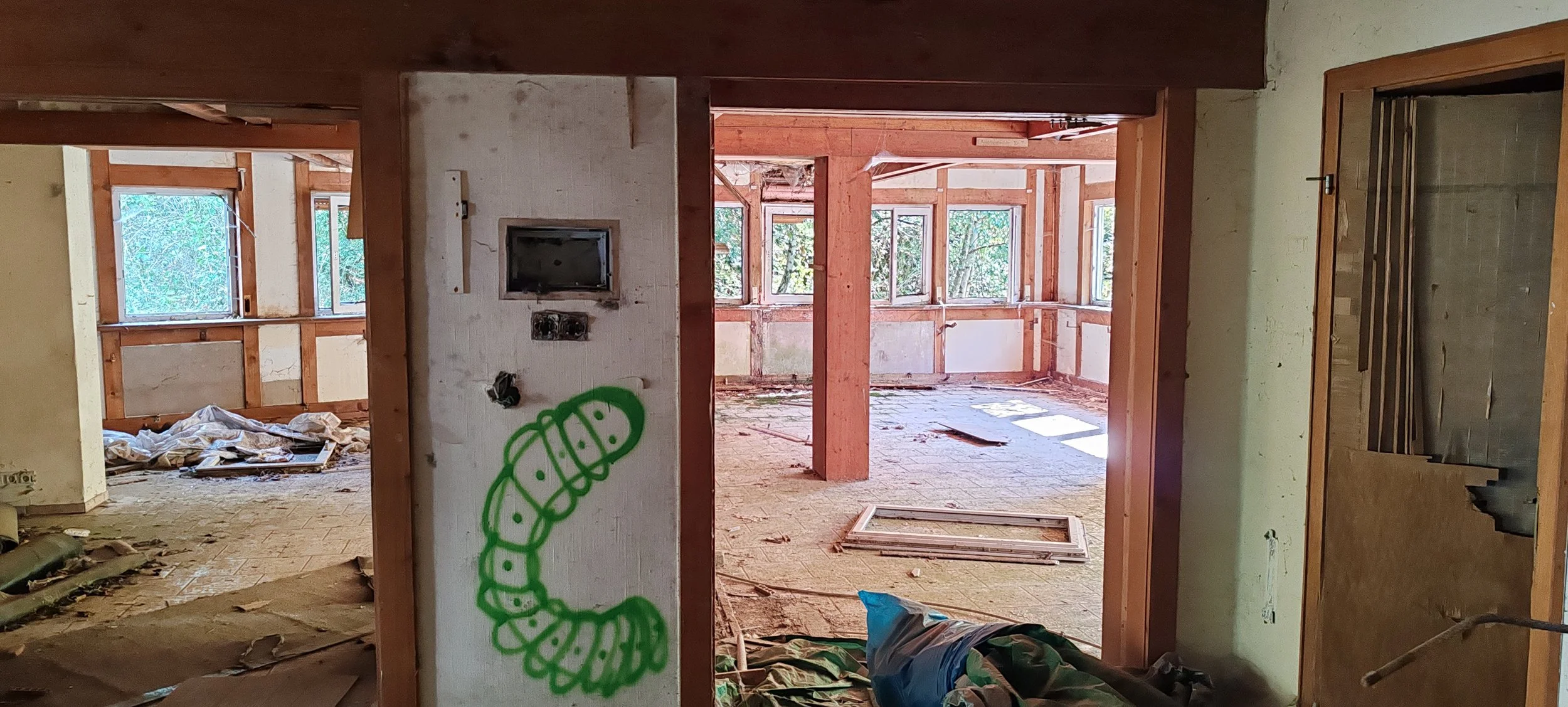 Innenansicht eines alten, verlassenen Hauses mit unordentlichem Boden und offenen Fenstern, Holzrahmen und Graffiti auf einer weißen Wand.