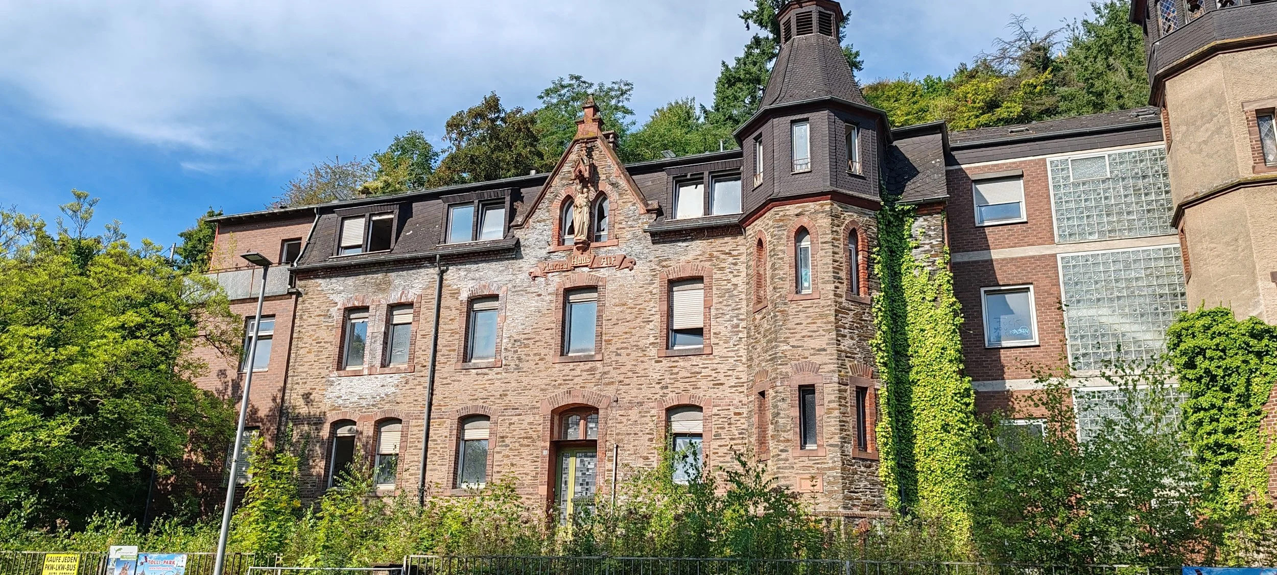 Historisches, mehrstöckiges Gebäude mit auffälligem Turm und Verzierungen auf der Fassade, umgeben von grünen Bäumen und Pflanzen, bei sonnigem Himmel.
