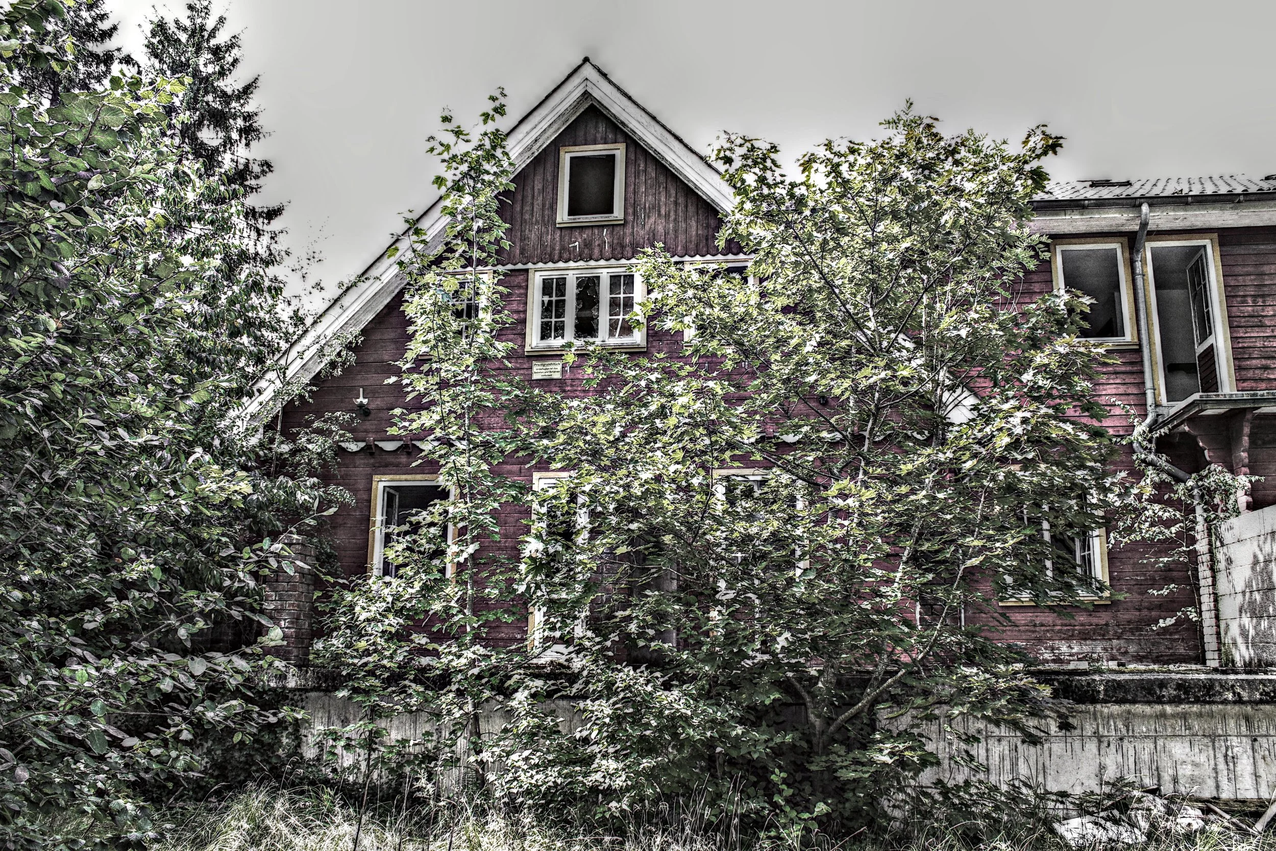 Ein altes, rotes Holzhäuschen mit weiß umrandeten Fenstern, teilweise von Bäumen und Sträuchern verdeckt, auf einem steilen Hang an einem bewölkten Tag.