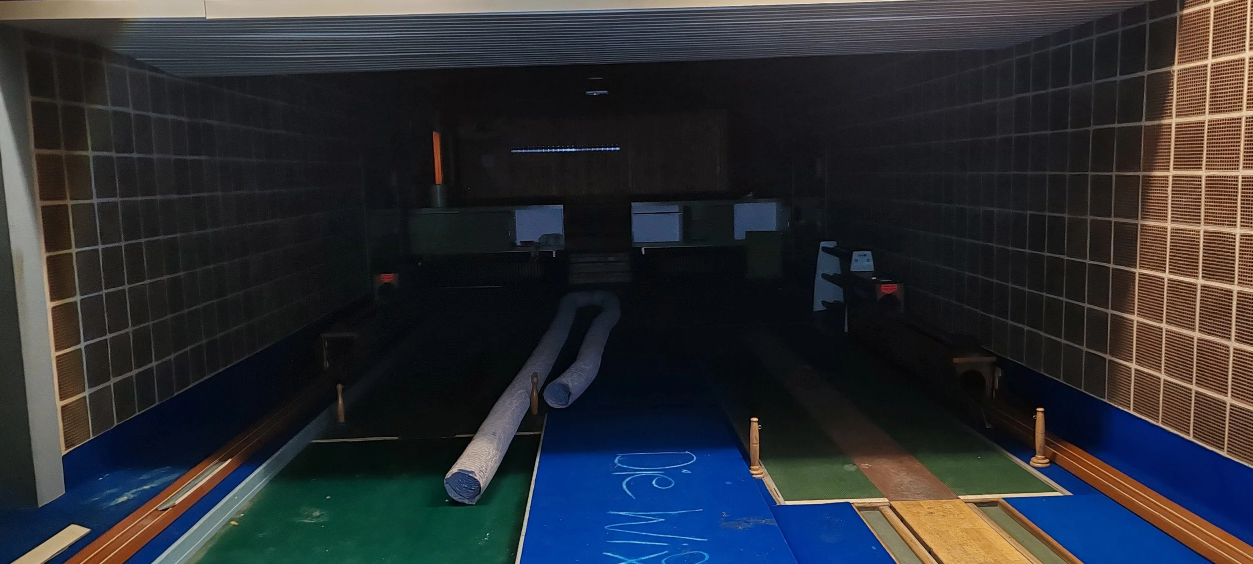 Ein dunkler, leerer Raum mit einem Minigolfplatz, grünen und blauen Spielfeldern, Röhren und Hindernissen, beleuchtet nur schwach.
