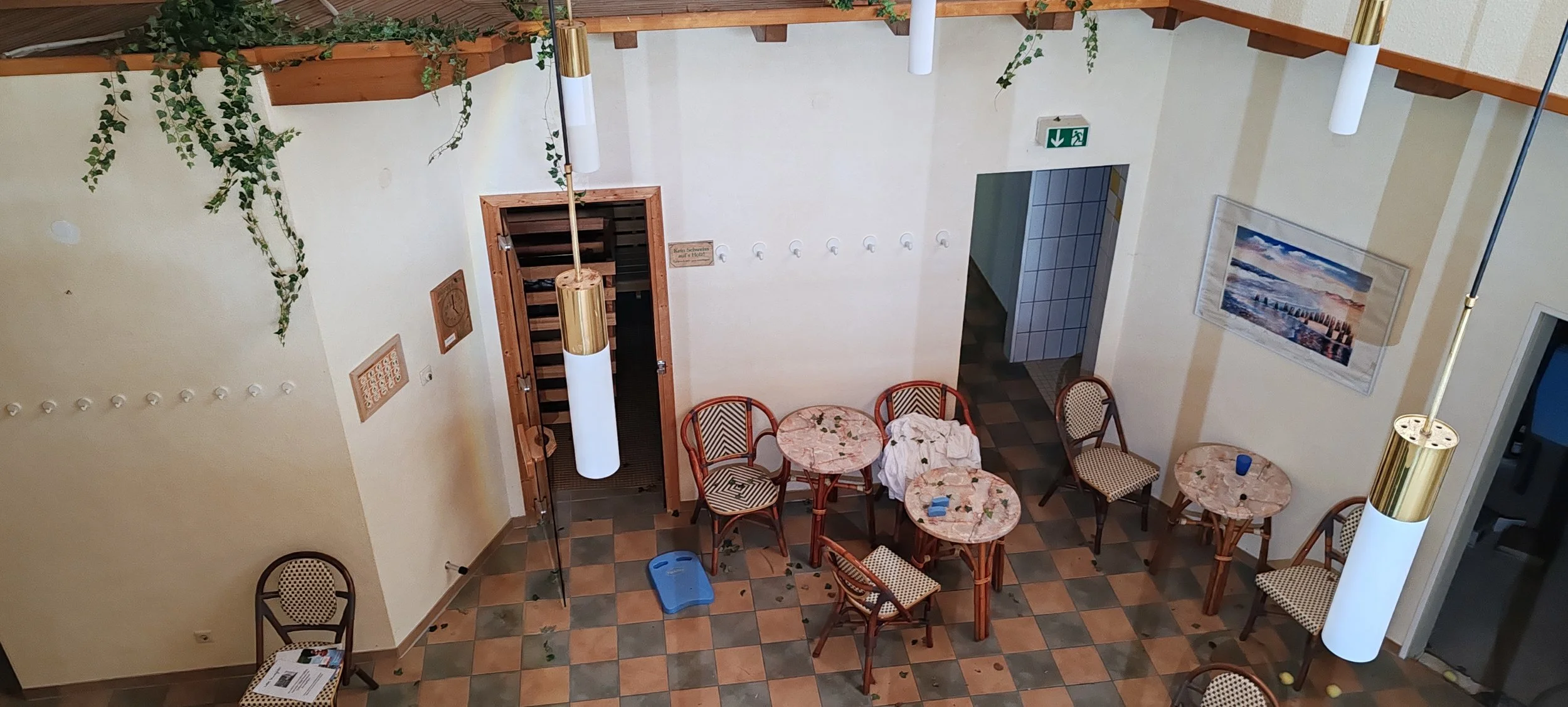 Mehrere Tische und Stühle in einem hell erleuchteten Raum, teilweise mit Gebüsch und Pflanzen an der Decke, Wanddekoration inklusive einem großen Bild und mehreren Haken an der Wand, auf dem Boden liegen ein paar Papierstücke und ein blauer Fußhocker