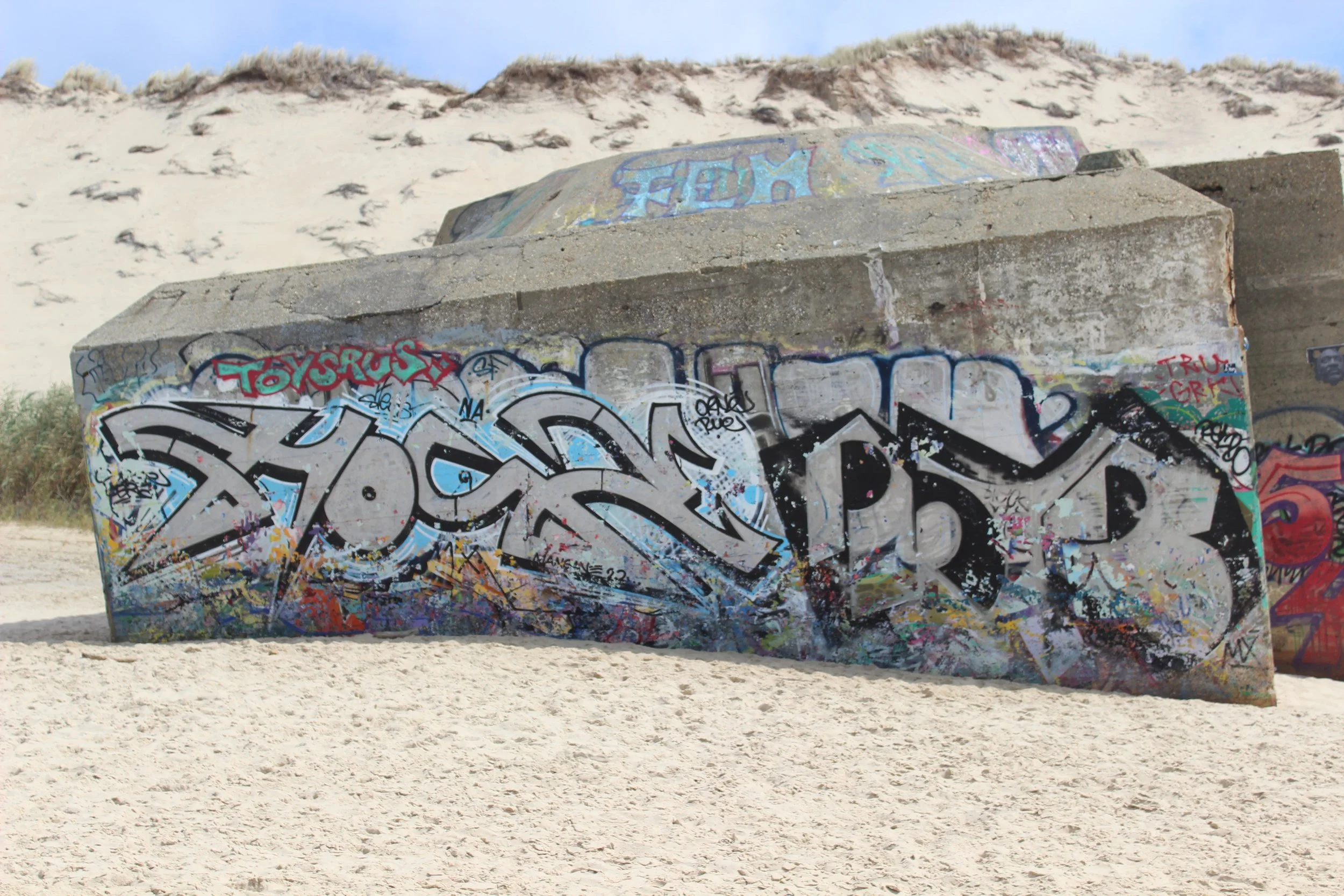 Graffiti-Kunst auf einem Betonsockel am Strand mit Sand und Hügel im Hintergrund.