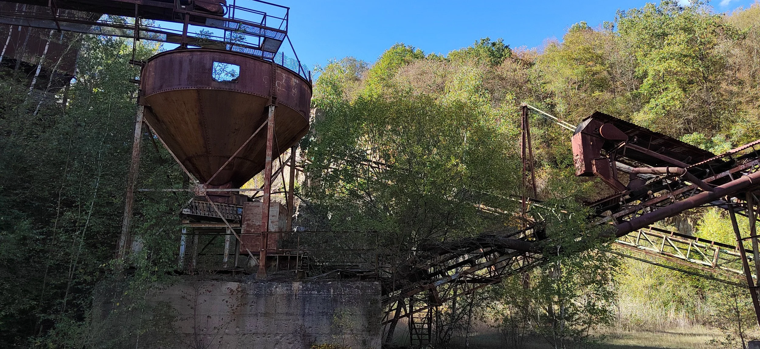 Verrostete alte Industrieanlagen im Wald, bestehend aus großen Silos und Förderbändern, umgeben von Bäumen und strahlendem blauen Himmel.
