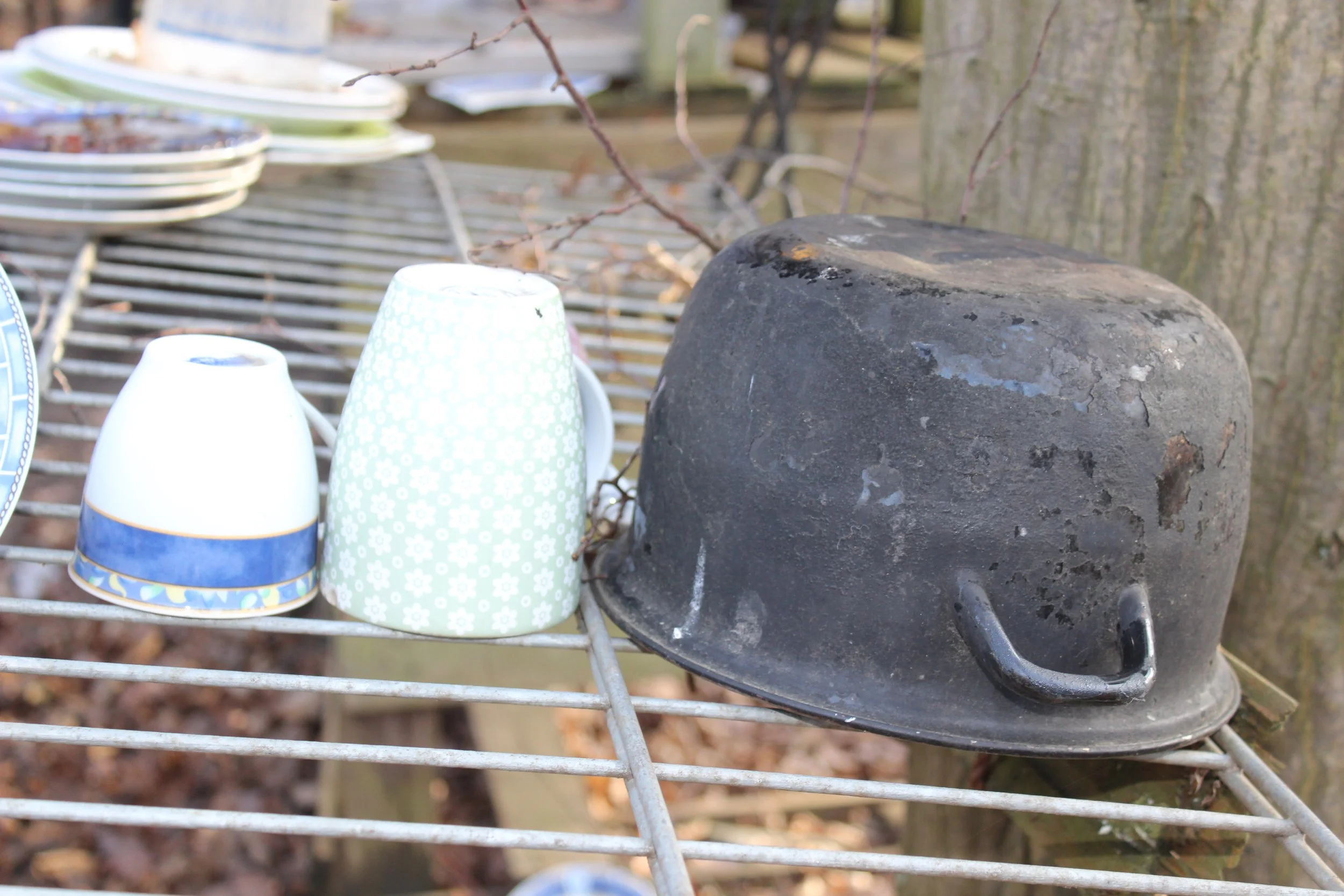 Ein alter, verrosteter Topf liegt auf einem Metallgitter, daneben befinden sich mehrere umgedrehte Porzellan-Tassen und Teller, im Hintergrund ein Baumstamm