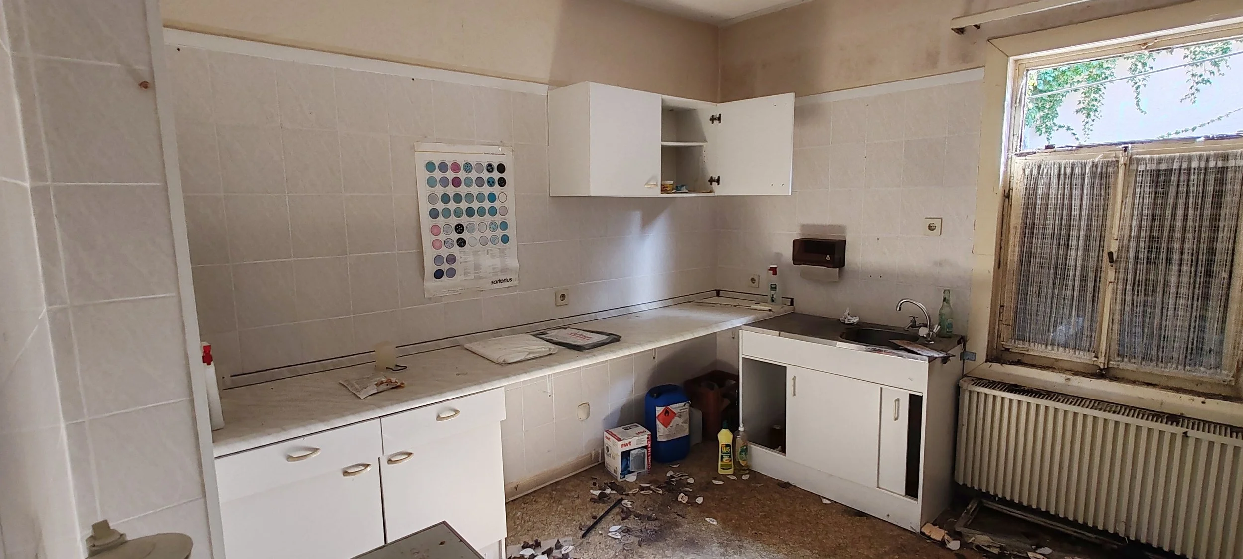 Ein verfallenes, unordentliches Küchenraum mit schmutziger Arbeitsfläche, offenem oberen Schrank, schmutzigen Boden und einem alten Fenster mit verschmutztem Vorhang.