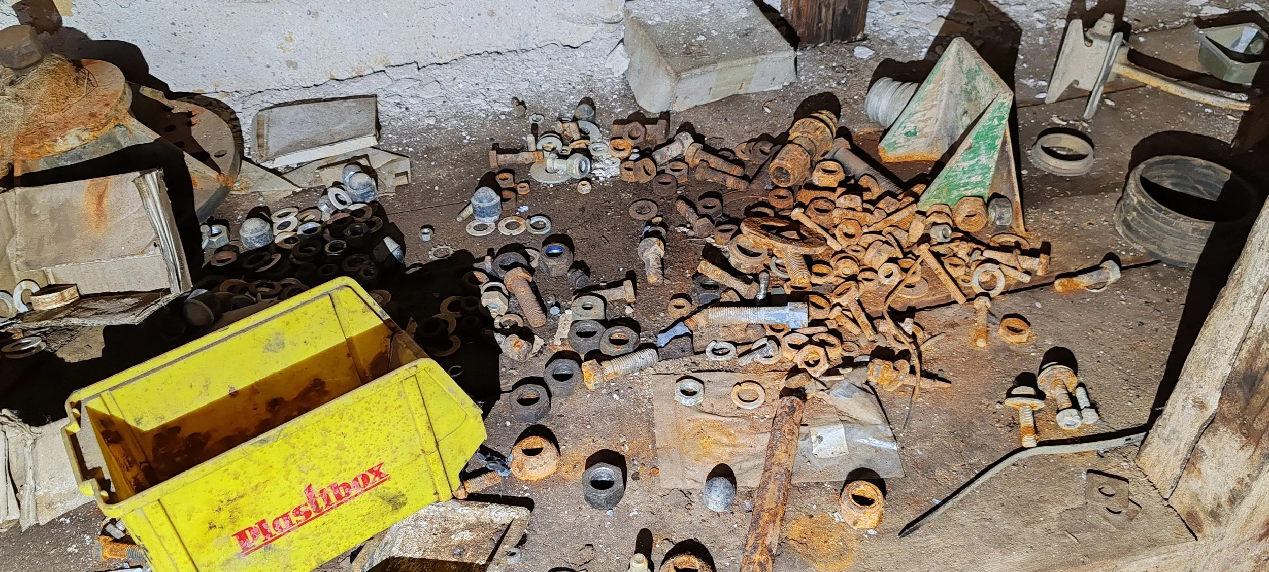 Boden mit verstreuten, alten und rostigen Schrauben, Muttern, Bolzen und rostigen Werkzeugteilen in einer Werkstatt oder Lagerraum, mit einer gelben Kiste und einigen Werkzeugen im Hintergrund.