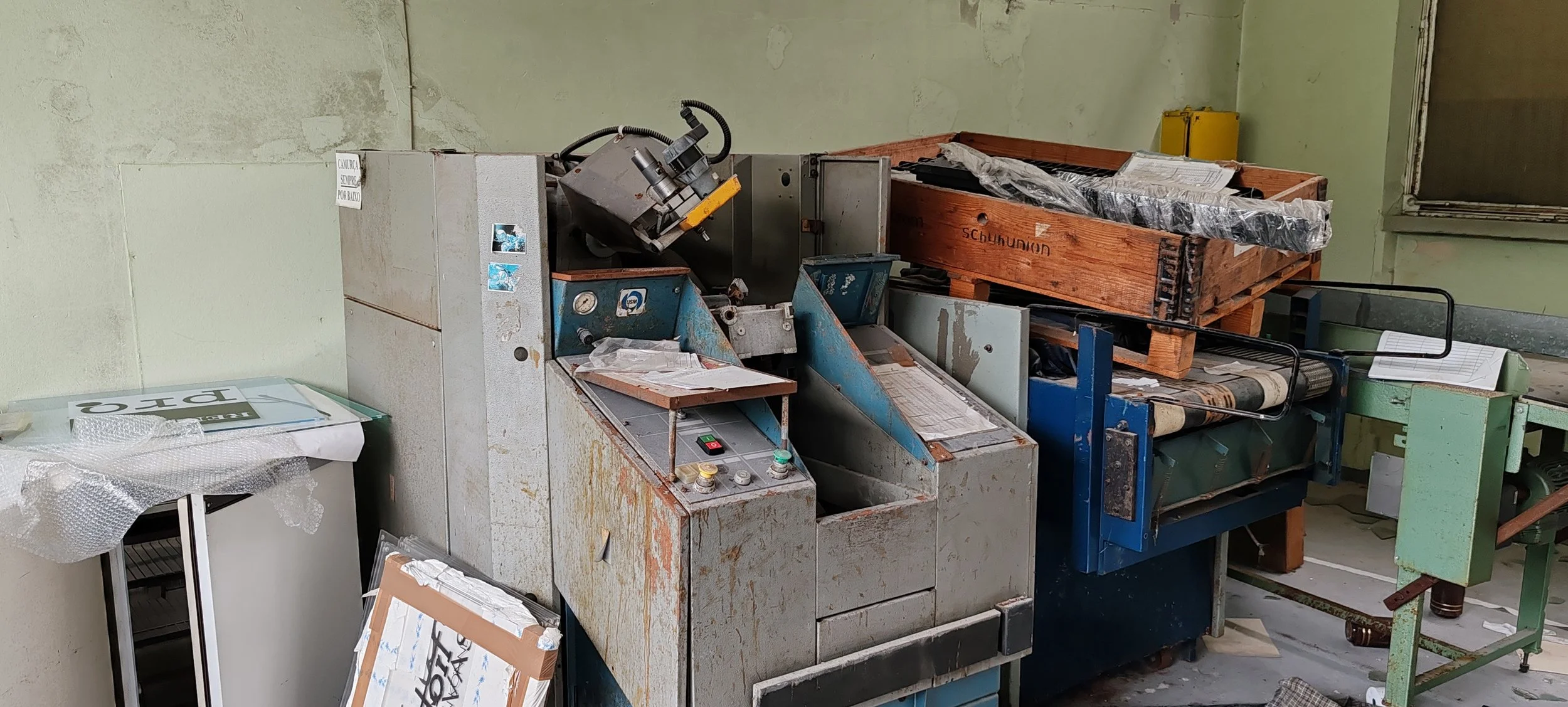 Altmaschine in einem veralteten, schmutzigen Raum, umgeben von Plastik, Papieren und Ersatzteilen, mit einem Holzkasten auf einem blauen Tisch.