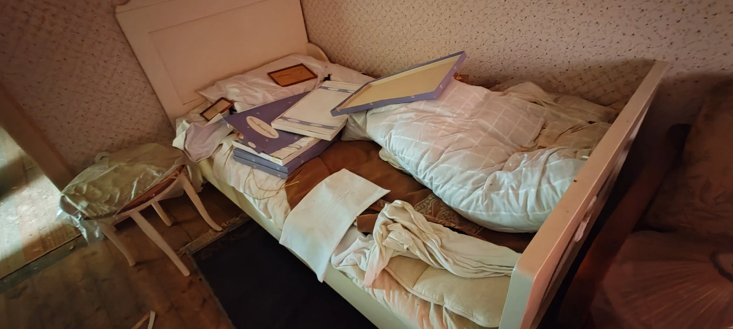 Ein unordentliches Bett mit zerzausten Bettwäsche, einem aufgeschlagenen Bilderrahmen, einigen Büchern, Papier und einem Plastiktüten-ähnlichen Gegenstand neben einem kleinen Nachttisch.