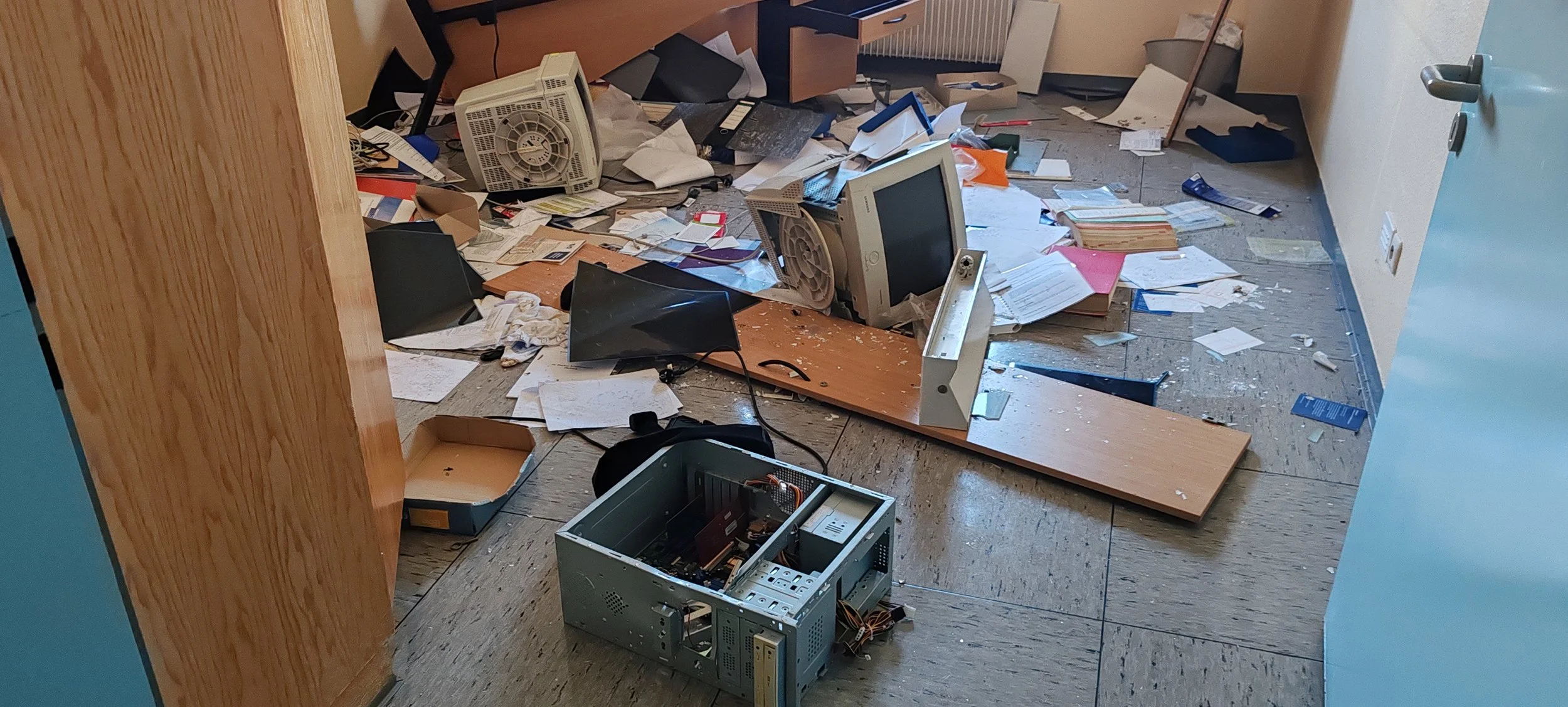 Ein beschädigtes Büro mit zerbrochenen Computerbildschirmen, einem geöffneten Computergehäuse, Papieren und anderem Müll auf dem Boden.