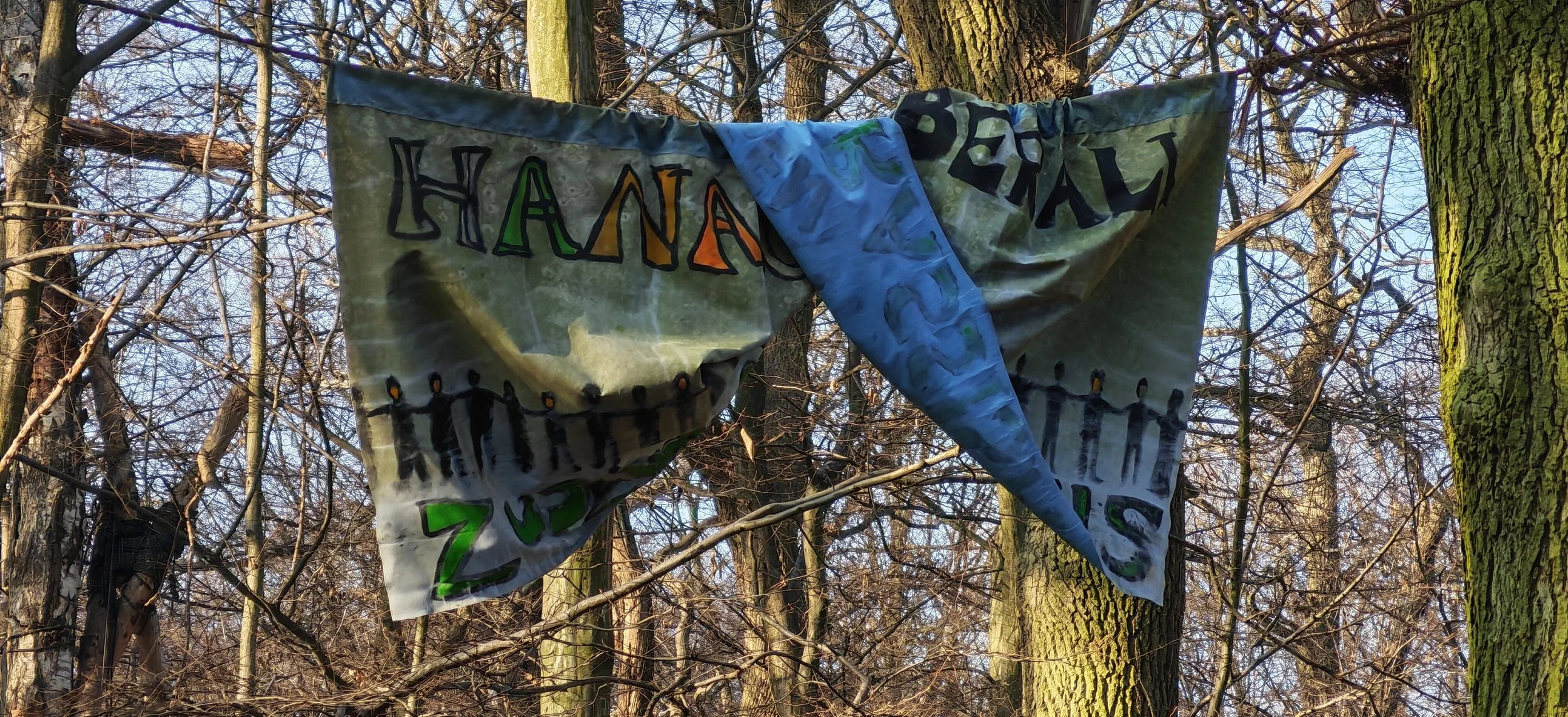 Bunte, handgemalte Flagge mit den Worten 'Hanna' und 'Zukunft' im Wald, zwischen Bäumen aufgehängt.