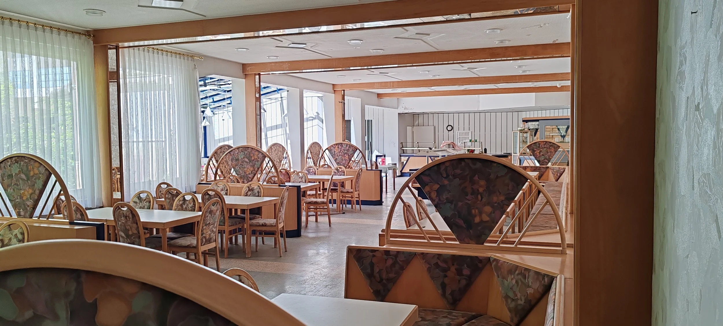 Leeres Restaurant mit Tischen und Stühlen, große Fenster mit weißen Vorhängen, Holzdecke mit Einbaulampen, im Hintergrund eine Theke und Küchenbereich.