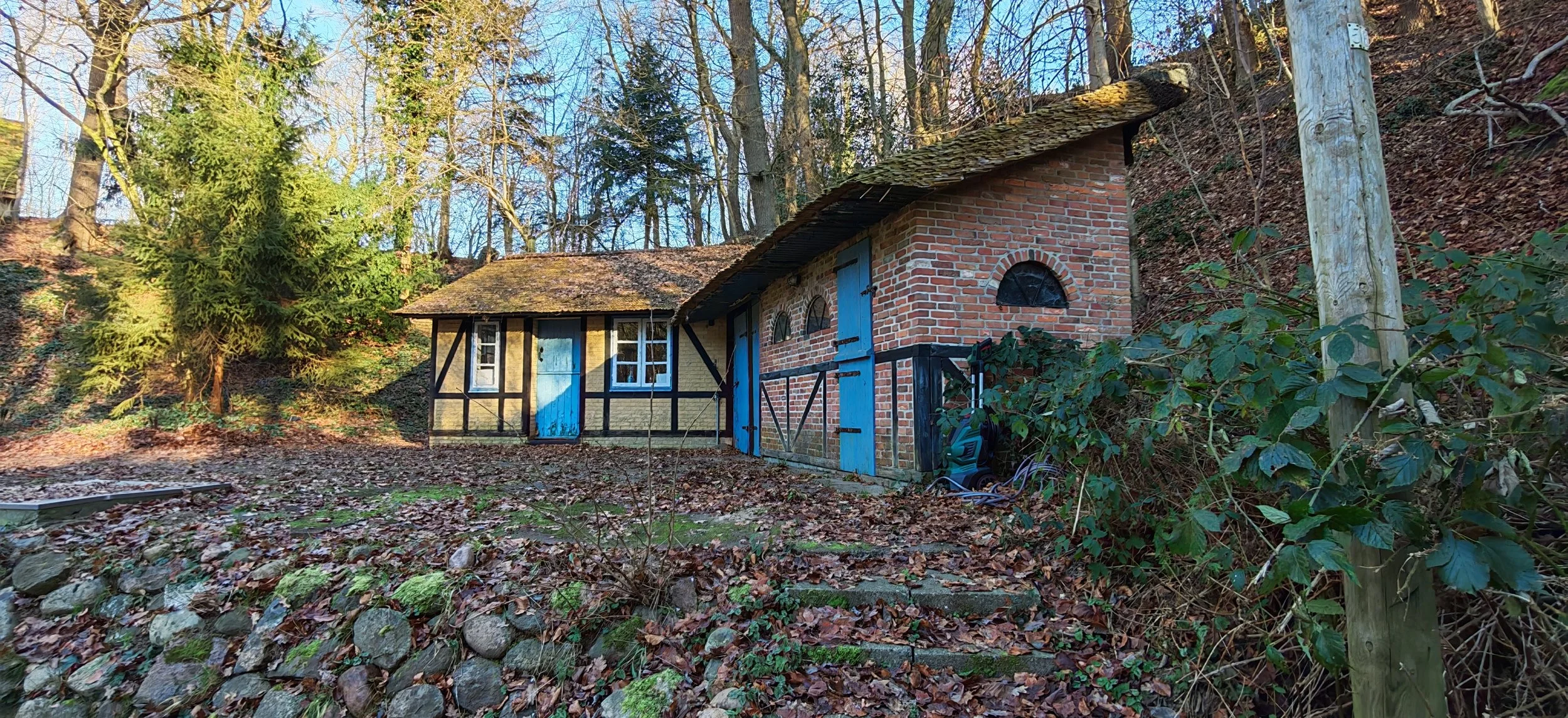Ein kleines Haus im Wald mit Backsteinmauer und blauen Türen. Es ist umgeben von Laub und Bäumen, im Vordergrund liegen Steine und Blätter.